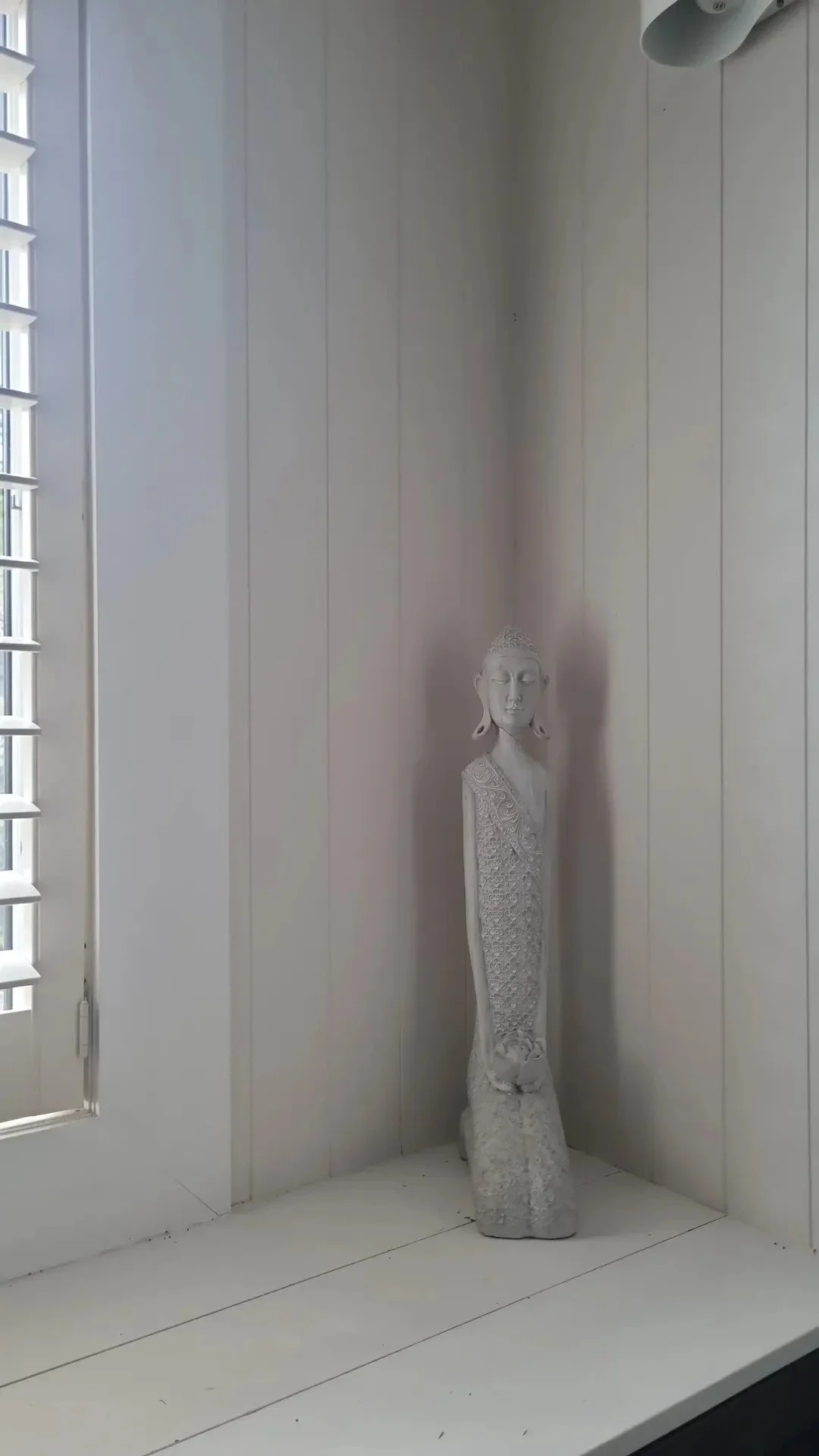 White Buddha statue with closed eyes, standing on a textured base in a corner of a room with white panel walls and a window with shutters.