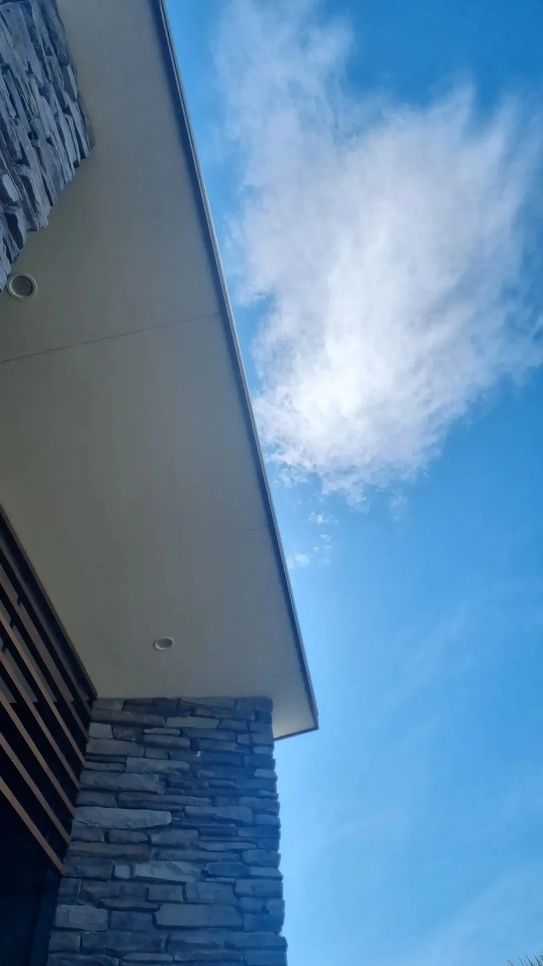 Part of a modern building with stone wall and overhang, against a bright blue sky with some clouds.