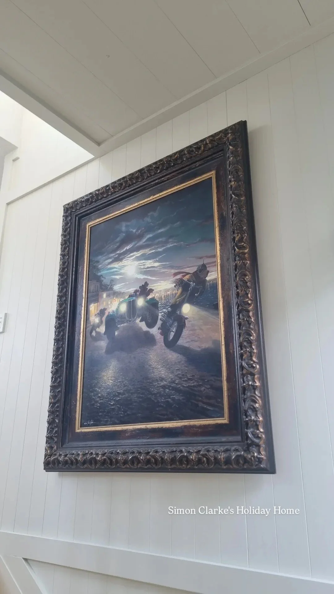 Painting of motorcyclists riding along a road at sunset, framed in an ornate black and gold frame, hung on a white wall.