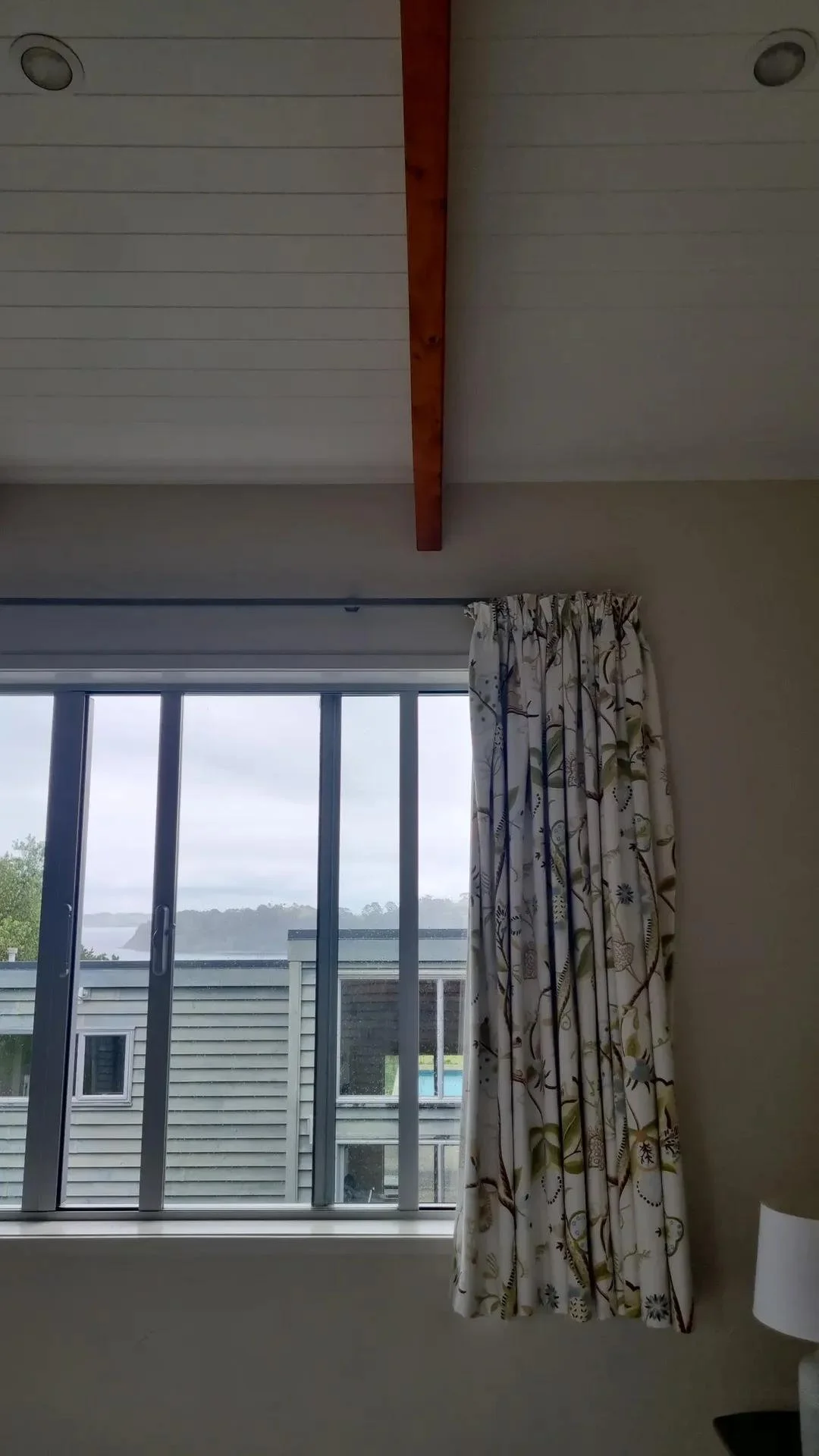 View through a sliding glass door showing a house and a lake or river outside, with a curtain on the right side of the door and a wooden beam on the ceiling.