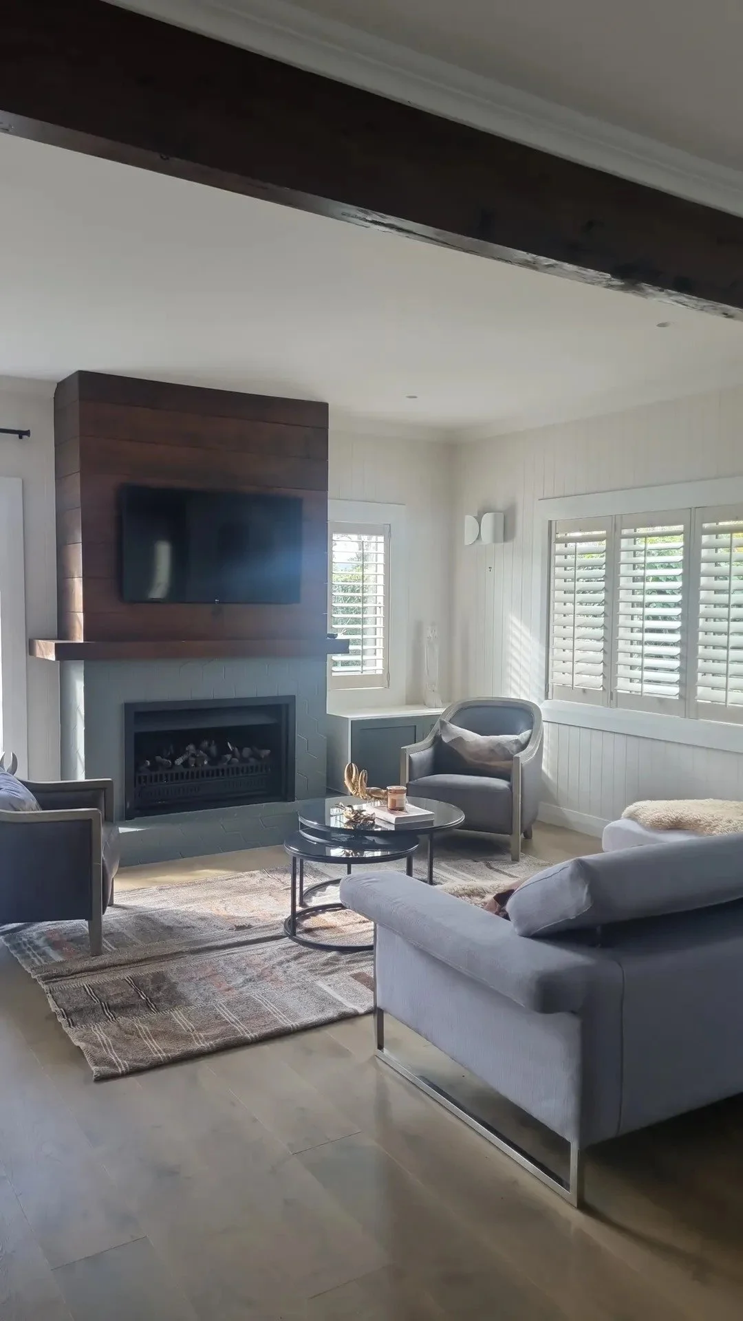 Living room with white walls, large windows with plantation shutters, a gray armchair, a white sofa, a black coffee table, a patterned area rug, a wall-mounted flat-screen TV, and a fireplace.