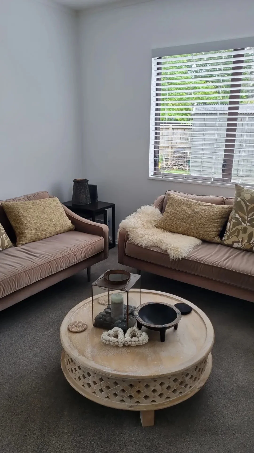 A cozy living room with two beige sofas, decorative pillows, a round coffee table, a window with white blinds, and a side table with a lamp. Outside the window, green trees and a small shed are visible.