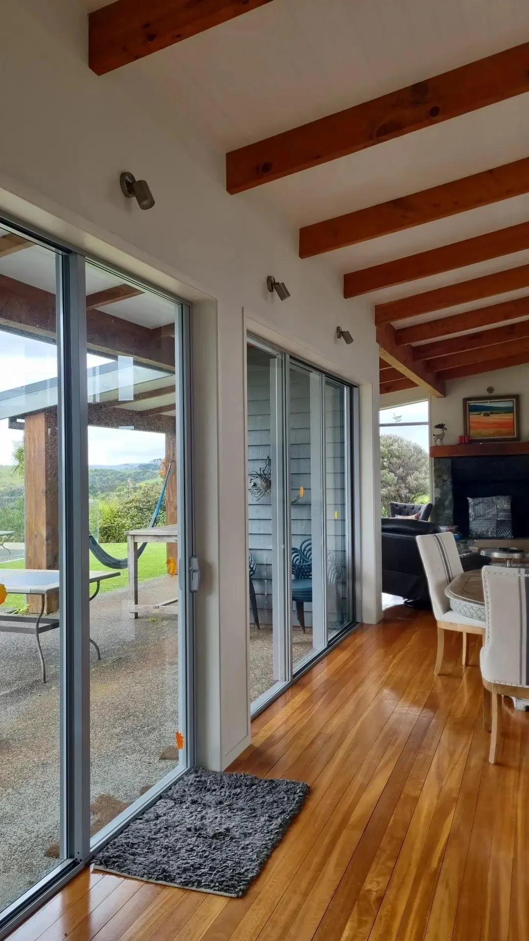 Inside view of a room with wooden flooring, sliding glass doors leading outside, and a ceiling with exposed wooden beams. Outside, there is a patio area with a hammock, a table, and chairs. Inside, there are chairs, a fireplace with artwork above it,