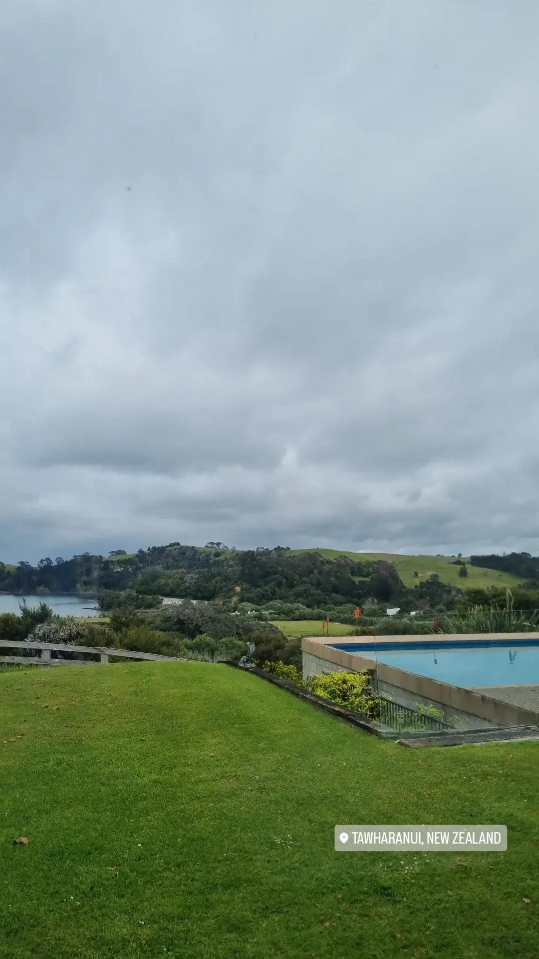 A lush green lawn with a swimming pool on the right, overlooking hilly terrain with green trees and a body of water, under a cloudy sky. Location tag indicates Tawharanui, New Zealand.