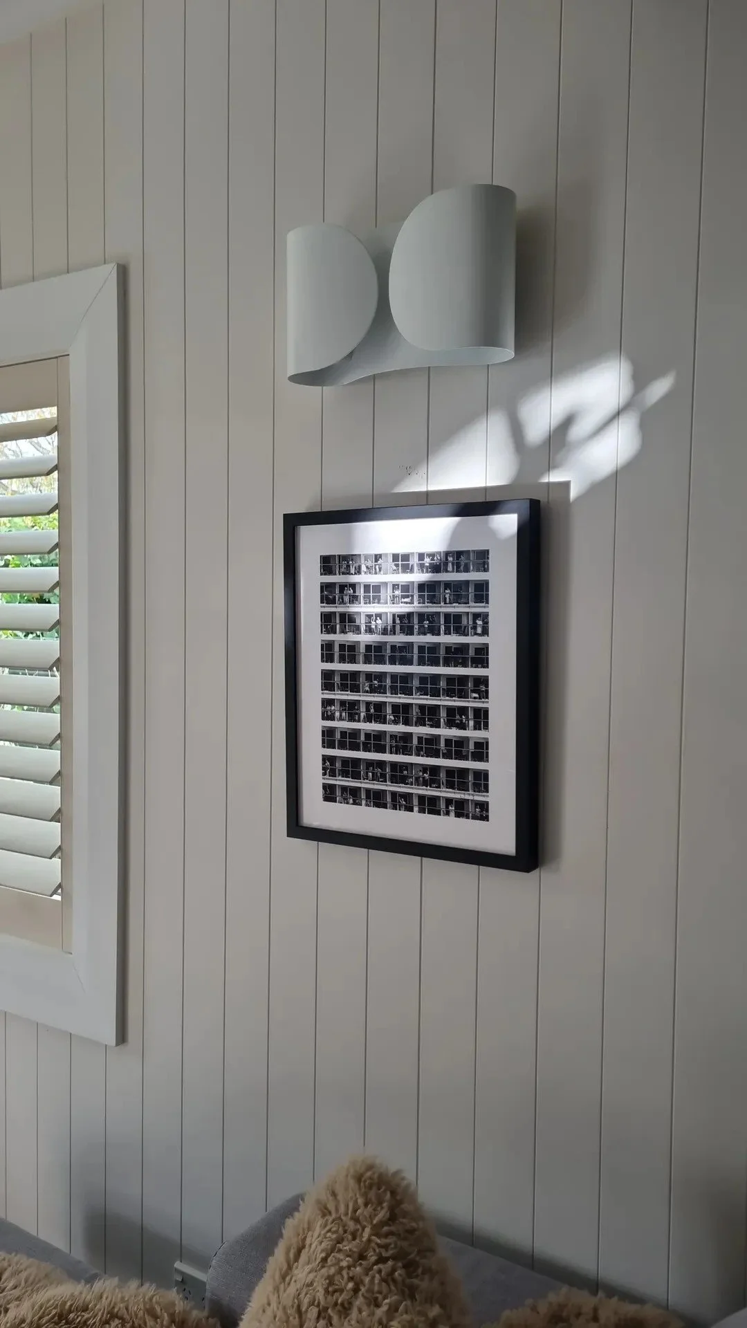 Interior of a room with white paneled walls, a framed black-and-white photo grid, a window with white shutters, and a ceiling-mounted abstract sculpture or art piece. Part of a couch with furry pillows is visible at the bottom.