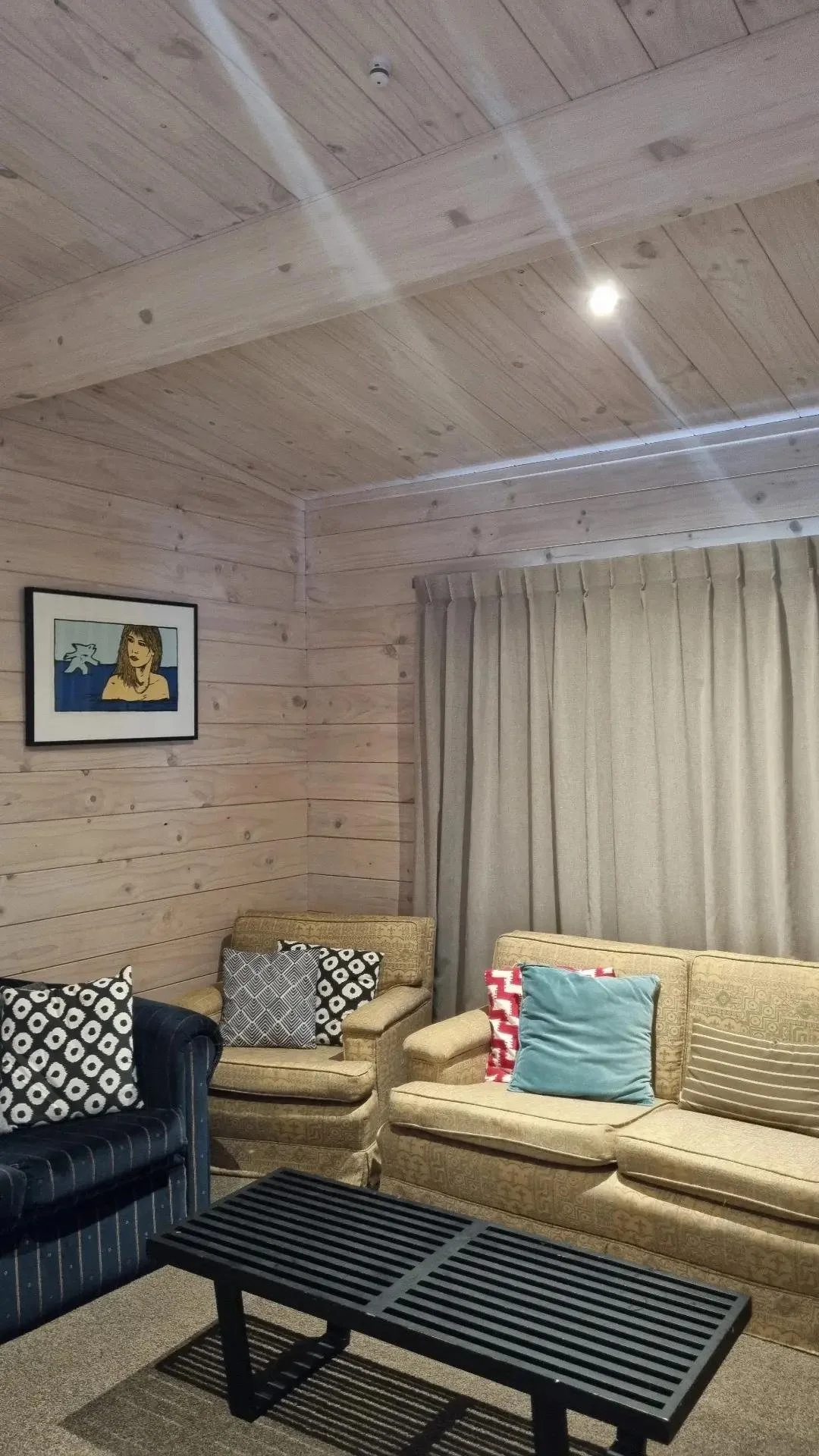 Living room with wooden paneled walls and ceiling, beige and navy blue sofas with patterned and solid cushions, a black slatted coffee table, a framed artwork of a woman, and beige curtains.