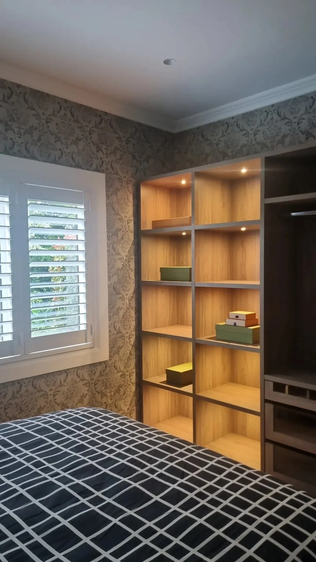 A bedroom corner with a window, a patterned wall, a wooden bookshelf with small storage boxes, and a bed with a grid-patterned black and white bedsheet.