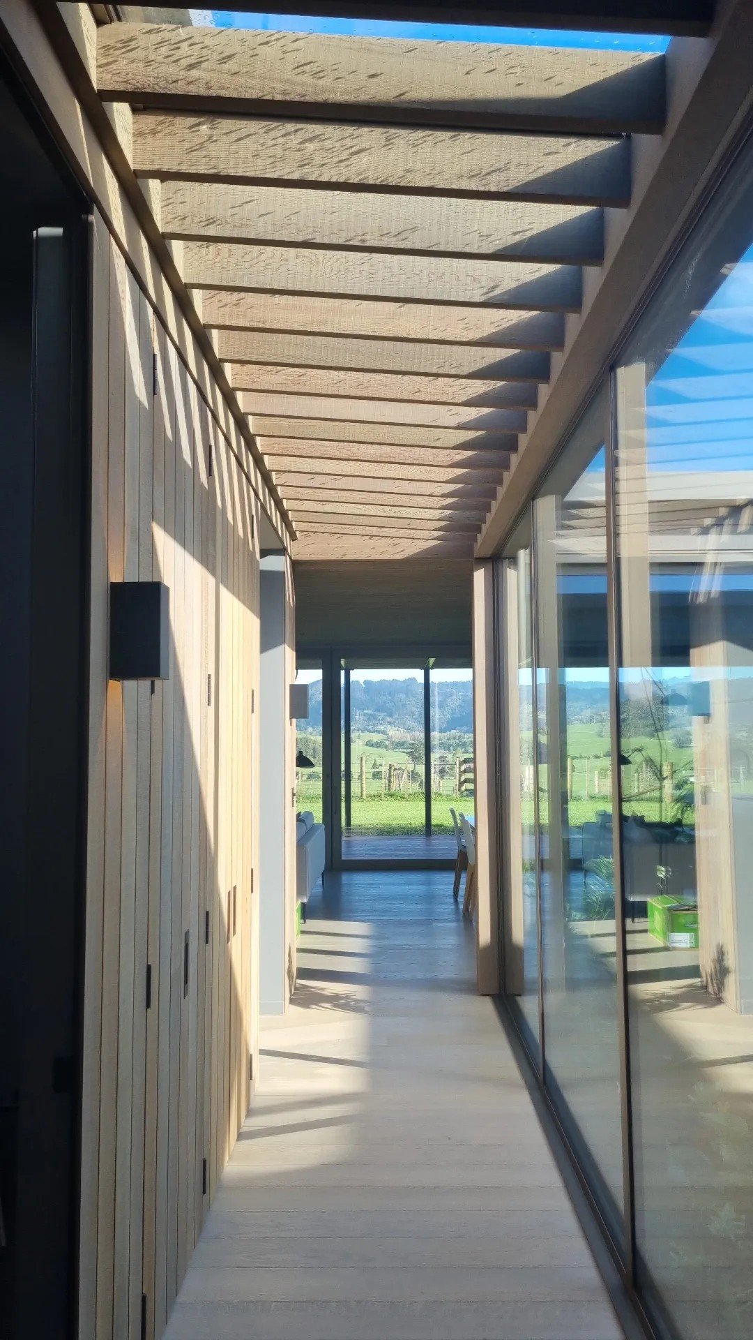 Long narrow balcony or corridor with wooden ceiling slats, glass sliding doors on the right, and a view of fields and hills outside.
