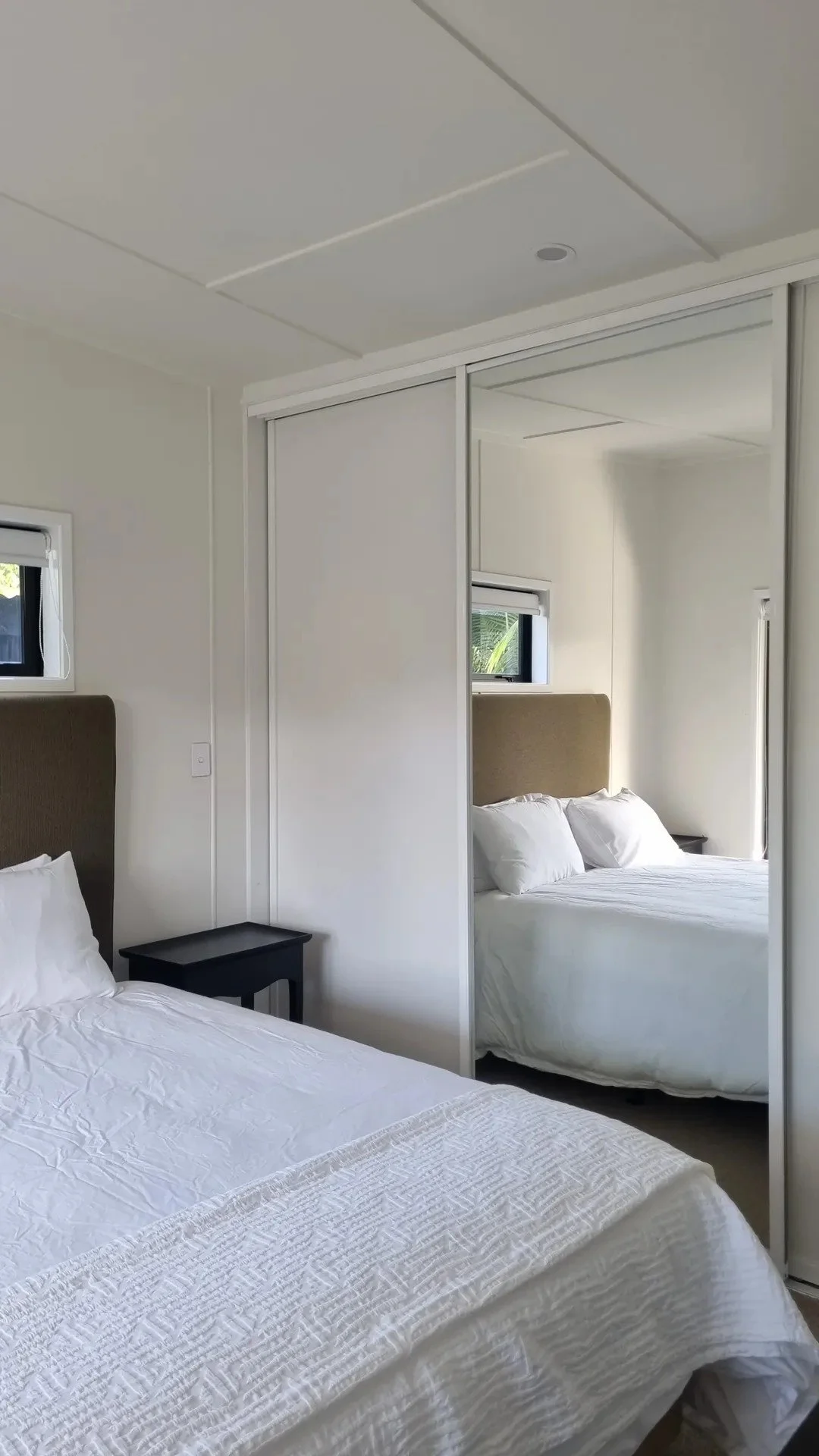 A bedroom with two beds, one in the foreground and one in the background, separated by a white sliding closet door with a mirror. The beds have white linens and pillows, and black nightstands are next to each bed. There are small windows with blinds 