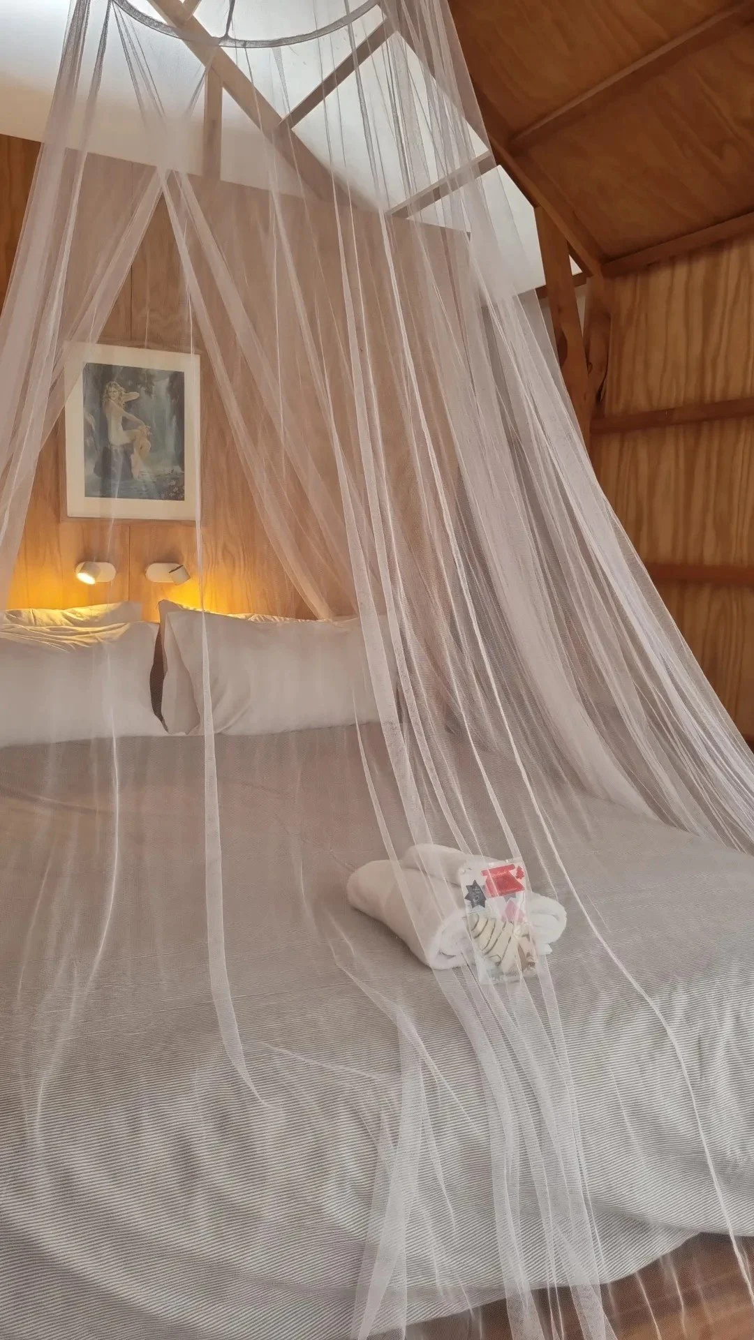 A neatly made bed with white sheets and a beige headboard, surrounded by a transparent white canopy. A towel with toiletries is placed on the bed, and there is a framed picture of a woman on the wooden-paneled wall above the bed with two small readin