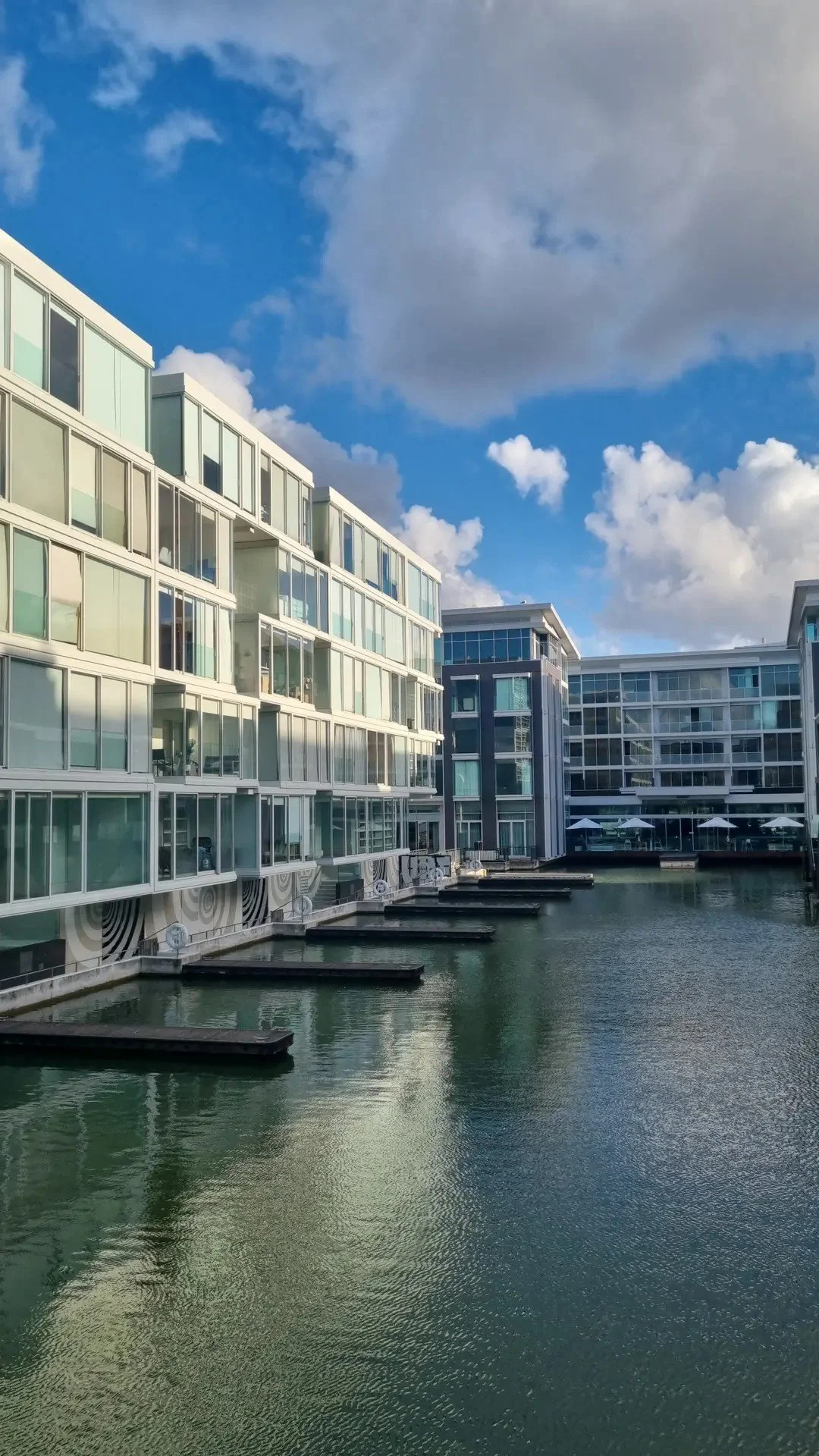Modern apartment buildings alongside a water canal, with clear blue sky and white clouds overhead.