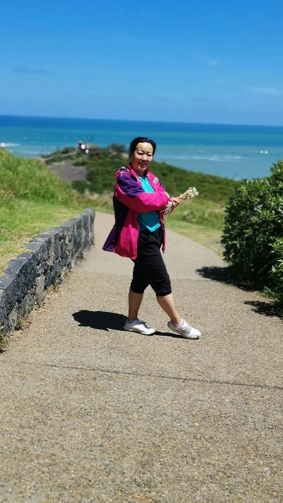 A woman in colorful outdoor clothing walking along a paved path near the ocean, holding a small bouquet of flowers, with blue sky and green shrubbery in the background.