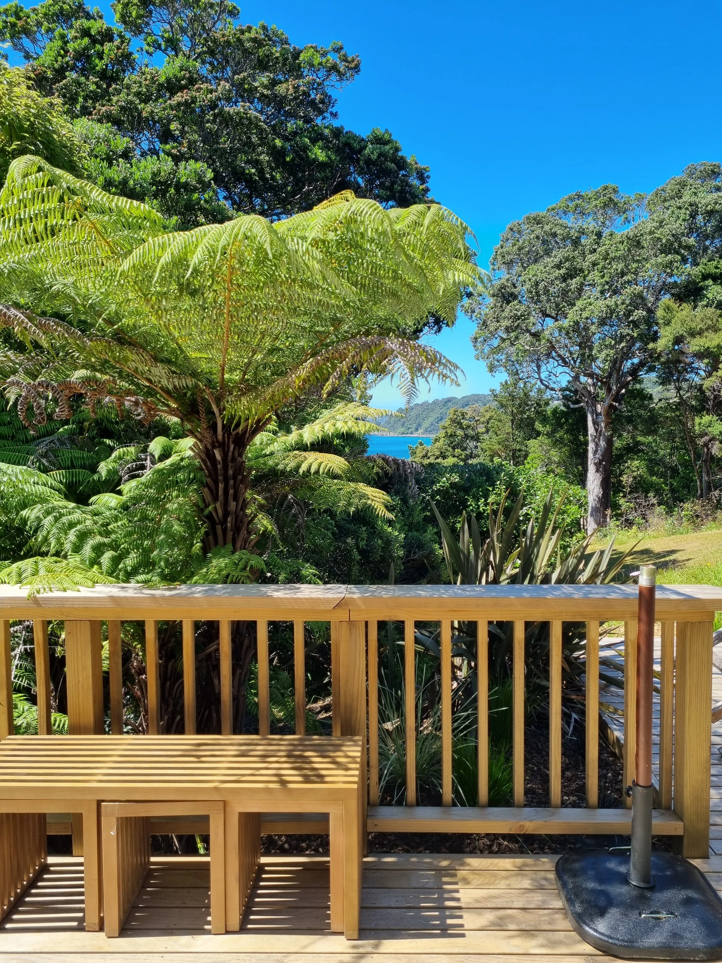 View of a wooden deck with a bench and a patio umbrella base, overlooking lush green trees, with a glimpse of water and distant hills under a clear blue sky.