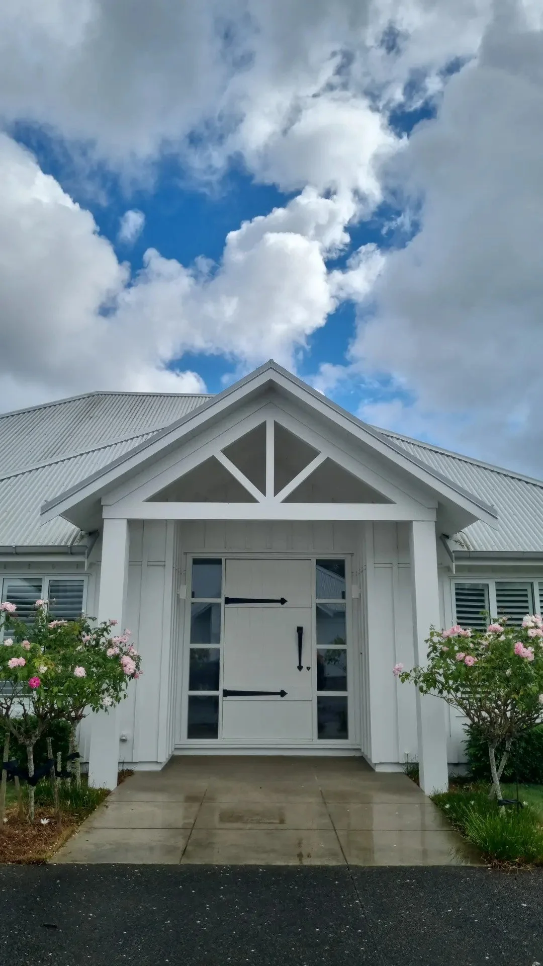 White house with barn-style door, surrounded by pink flowering bushes, under a blue sky with clouds.