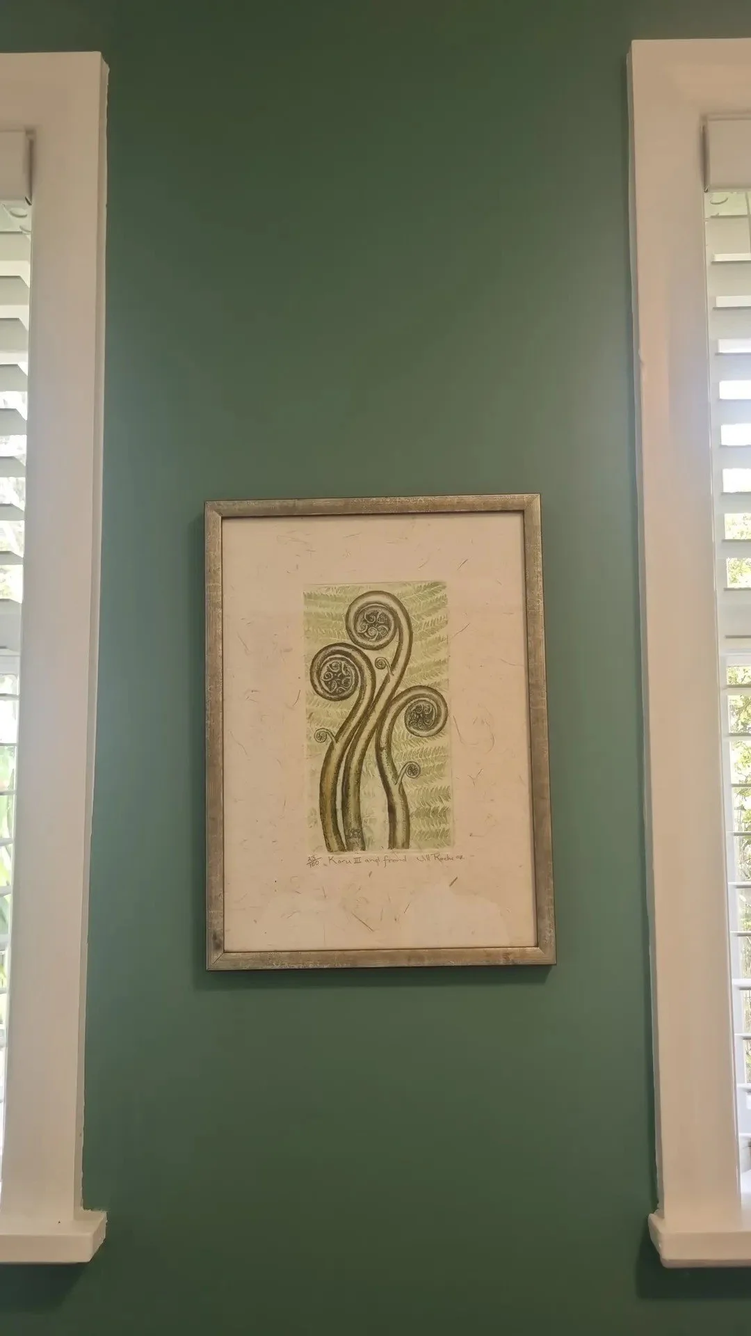 Framed artwork of a stylized tree with swirling branches on a green background, hanging on a teal wall between two white window shutters.