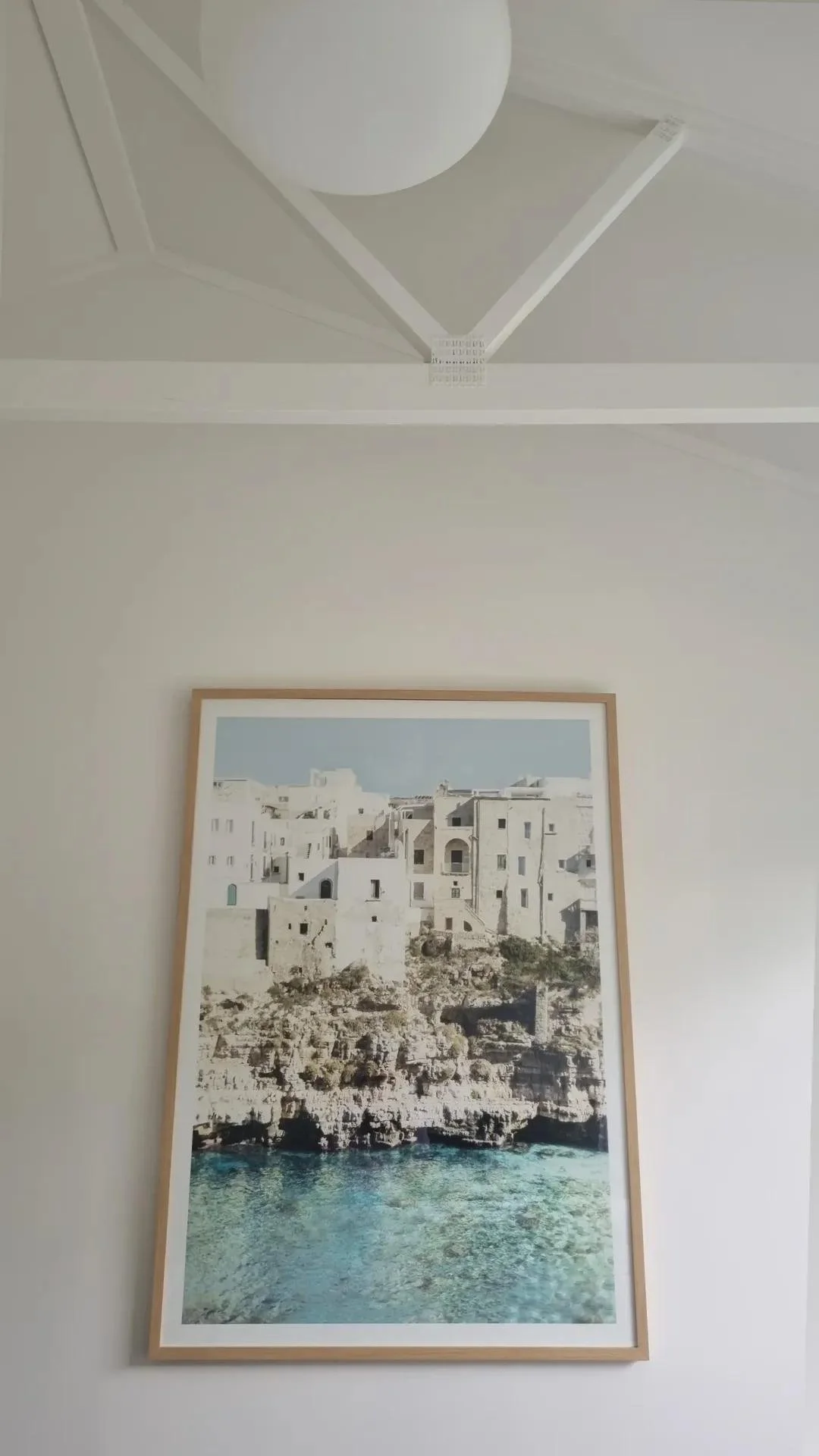 A framed picture of white buildings on a rocky coastline with clear blue water, hanging on a white wall.