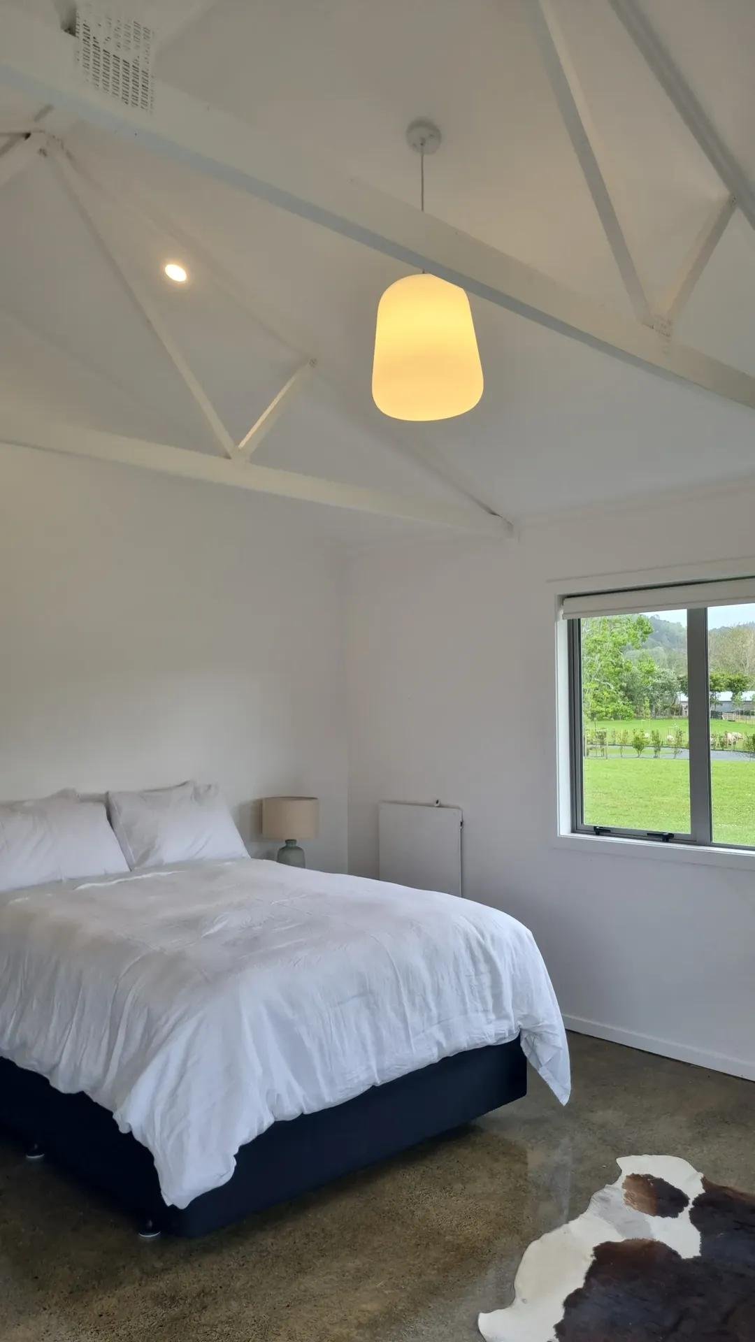 A bedroom with a white bed, white bedding, a nightstand with a lamp, a window with a view of green trees and grass, and a cowhide rug on a polished concrete floor.