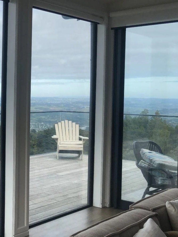 View through glass sliding doors of an outdoor wooden deck with a white Adirondack chair, overlooking a landscape of trees and distant fields under cloudy sky.