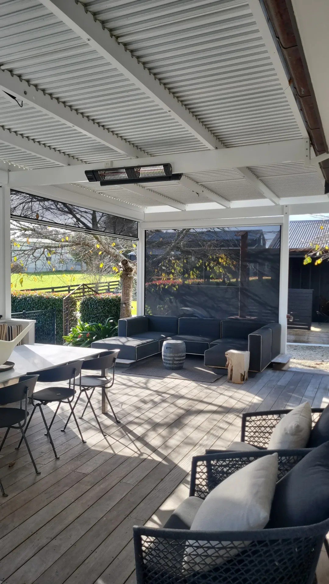A covered outdoor patio area with a modern black sectional sofa, a small decorative stool, and a tree visible outside through large glass windows. The patio has wooden decking and a mix of furniture including a dining table and chairs.