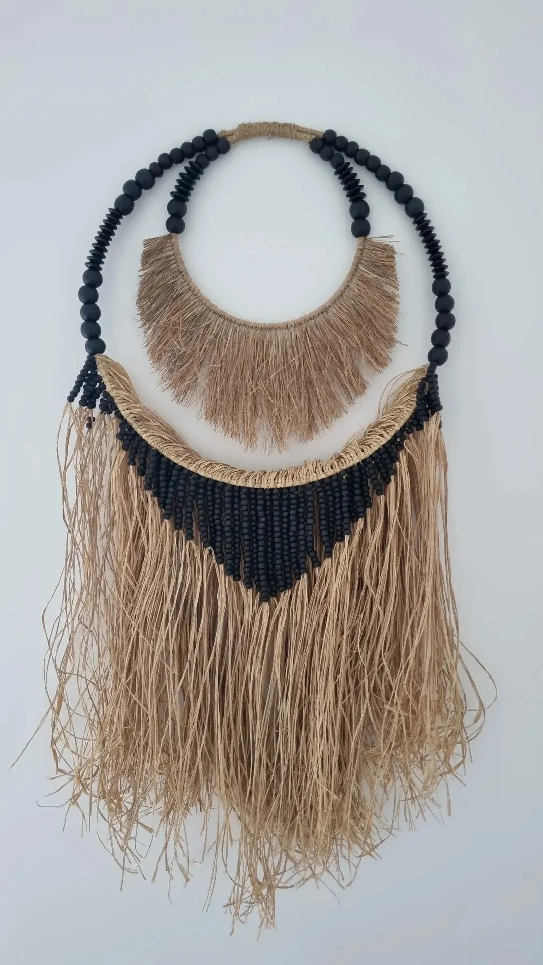 Two layered necklaces made of black beads and beige straw with fringe details, hanging against a plain white background.