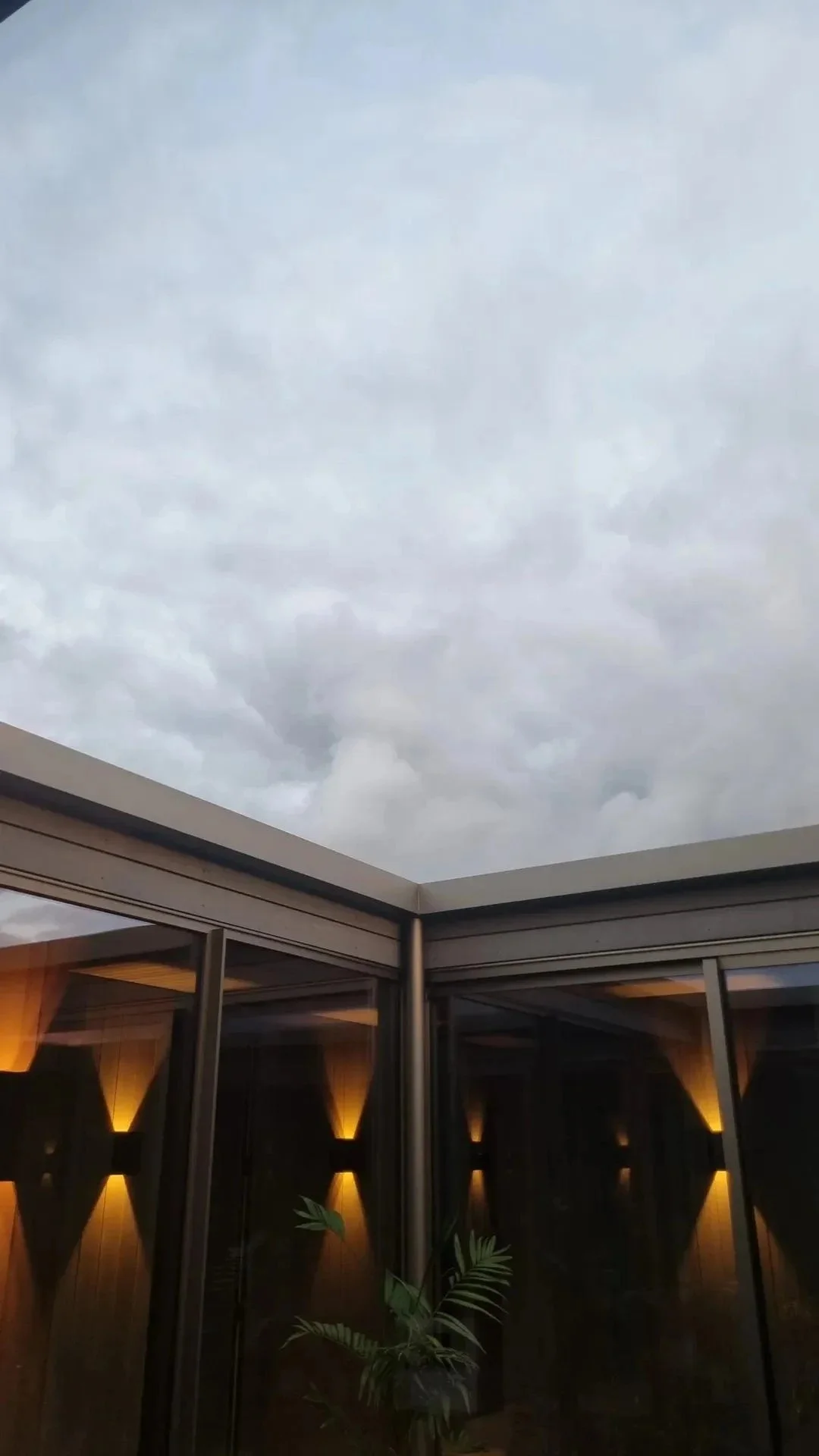 Photo of a cloudy sky above a modern building with glass walls and warm interior lighting, a potted plant is visible inside.