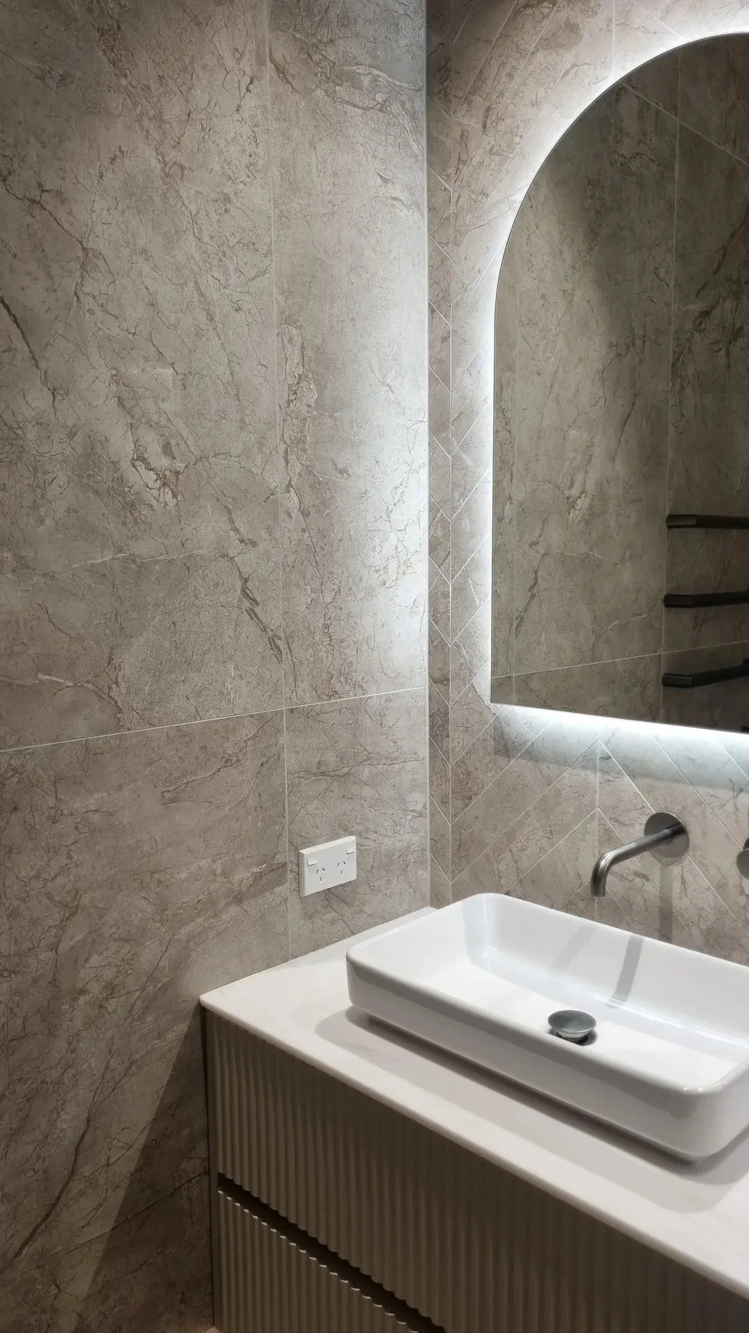 A modern bathroom with beige marble walls, a rectangular white sink, a large arched mirror with LED lighting, an electrical outlet, and dark towel racks.