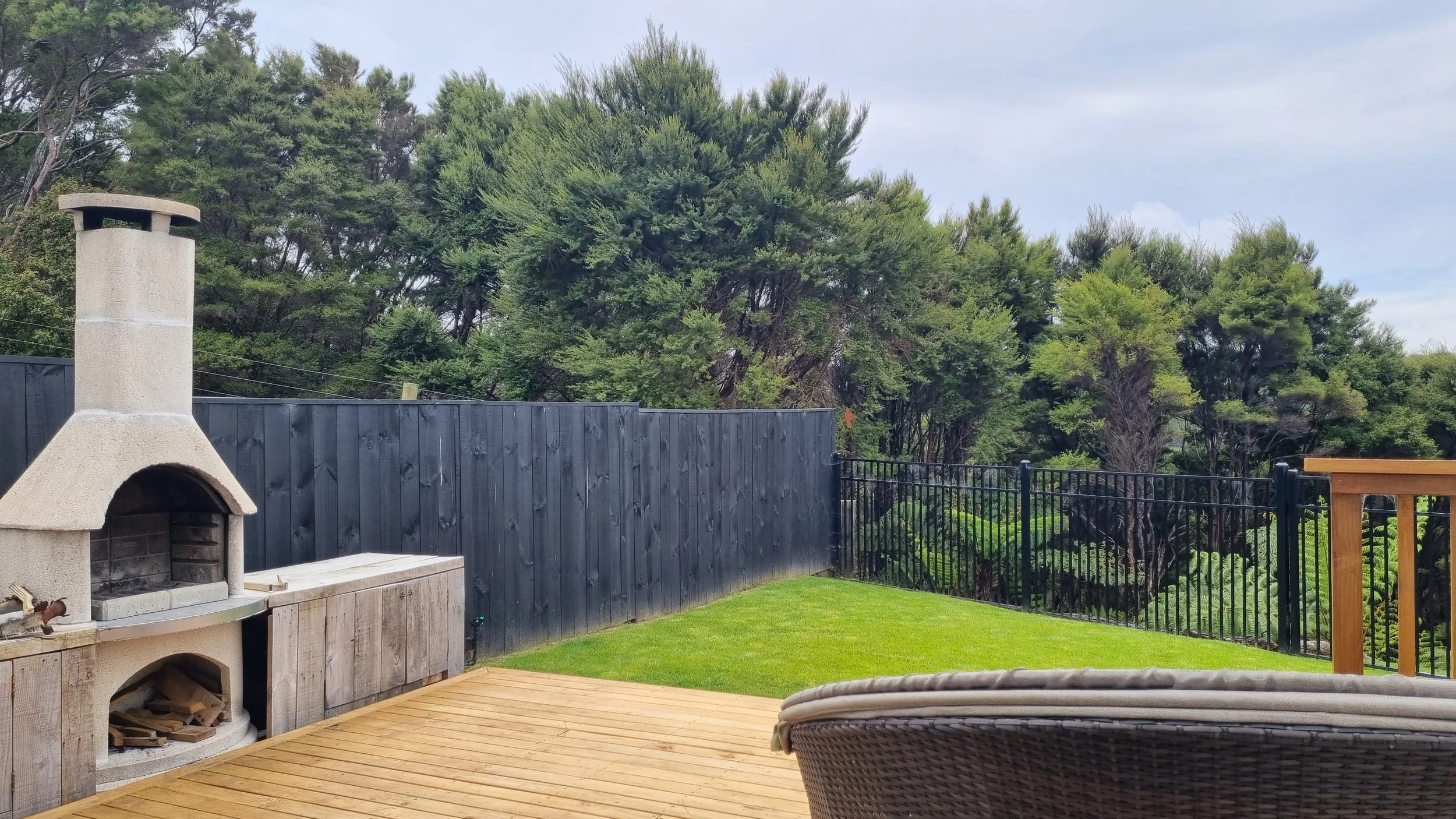 Backyard scene with a wood deck, a outdoor fireplace, a black wooden fence, a grassy area, and tall green trees in the background under a partly cloudy sky.