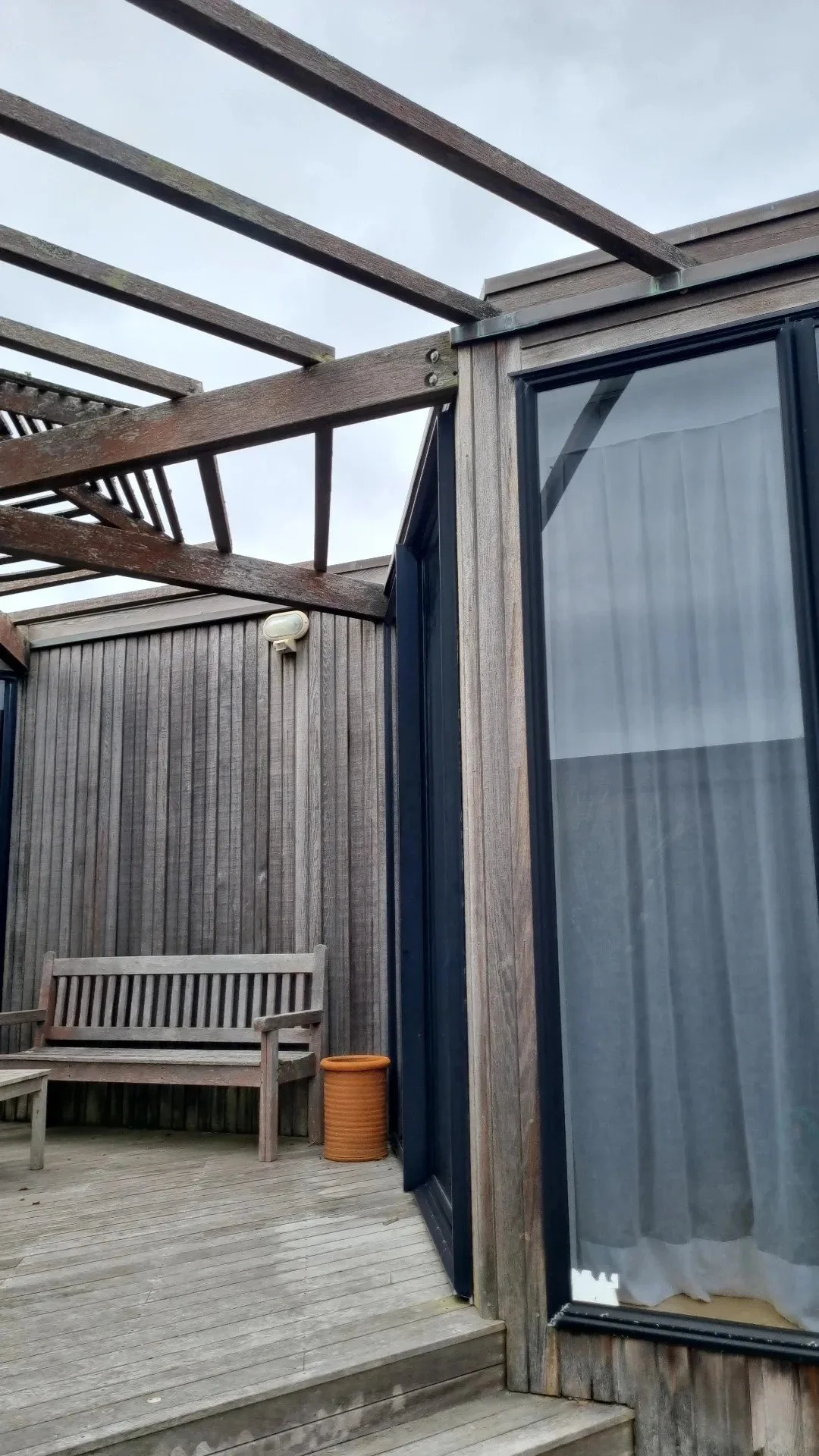 An outdoor wooden deck area with a wooden bench, a terracotta-colored planter, and a weathered wooden fence. Part of a house structure with large glass sliding doors and gray wooden exterior siding. Overhead, the pergola framework is incomplete with 