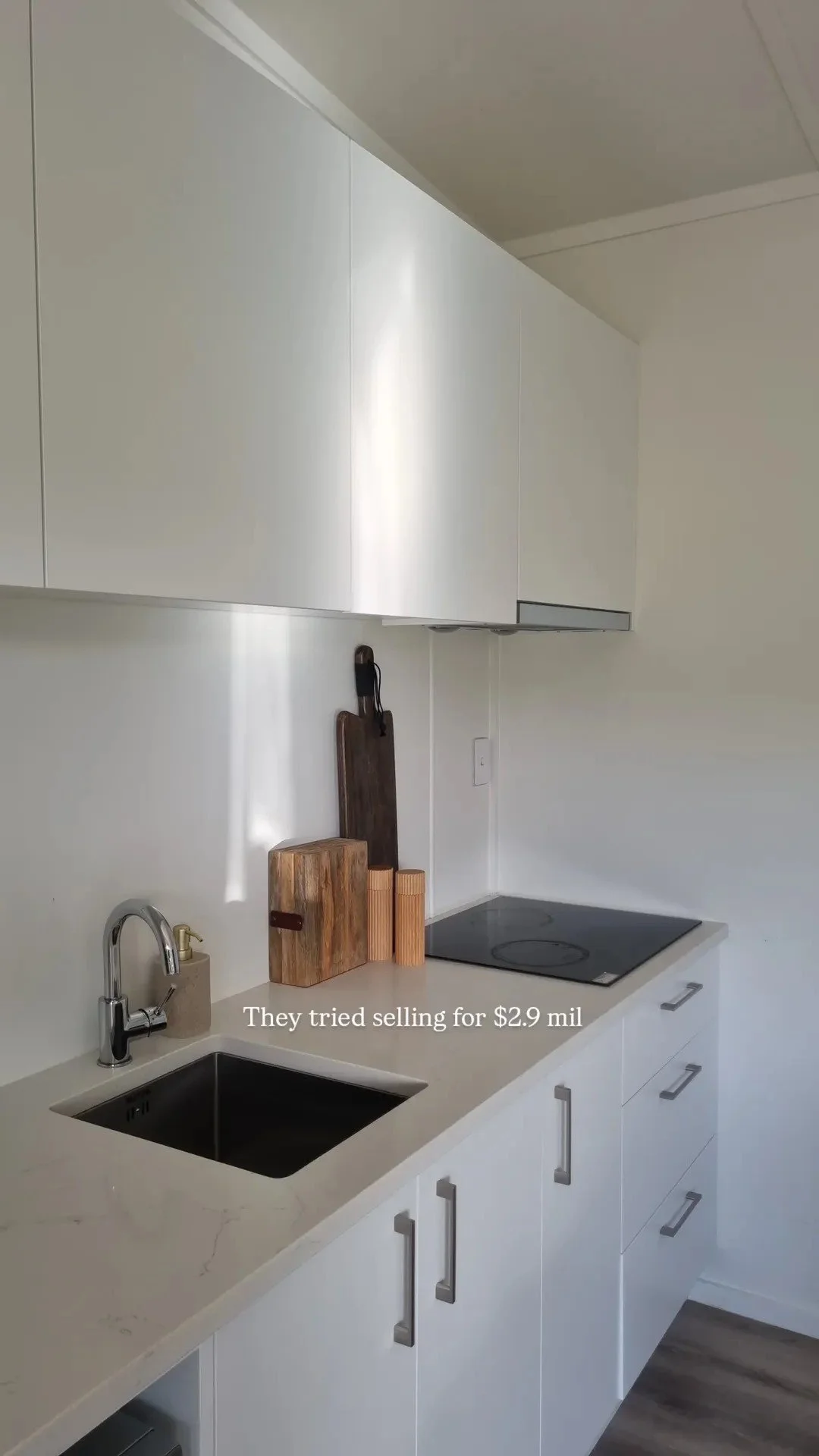 A modern kitchen with white cabinets, a black sink, a faucet, and a white countertop. Kitchen utensils including cutting boards and a soap dispenser are on the counter. There is a black electric stove and upper cabinets, with a wall outlet visible. T