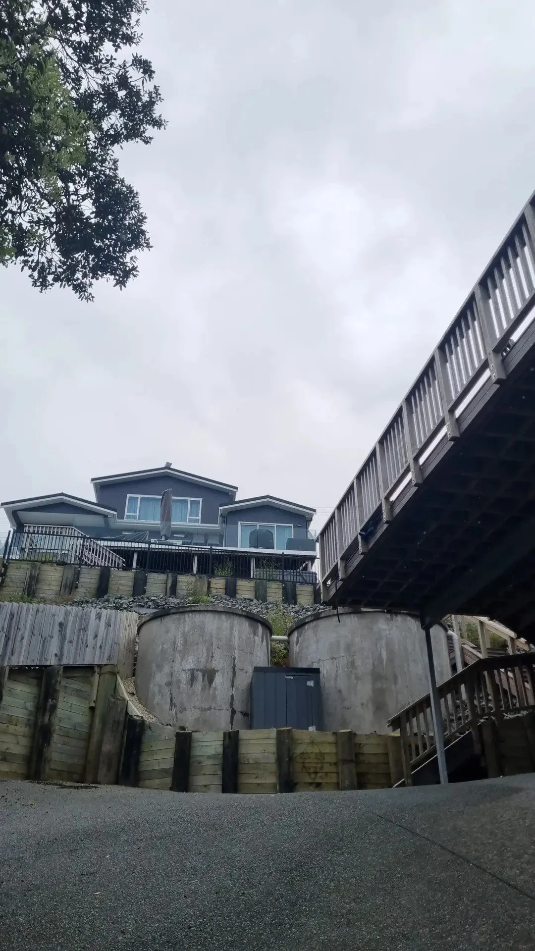 View of a house on a hill with a wooden balcony, surrounded by trees, under an overcast sky.