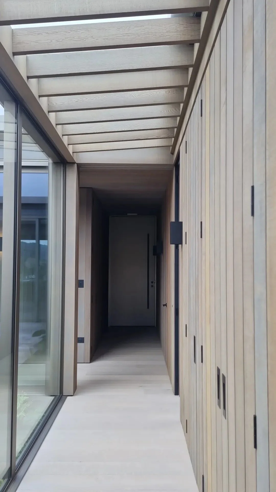 A modern hallway with wooden paneling on the right wall, glass sliding doors on the left, a ceiling with wooden beams, and a closed door at the end.