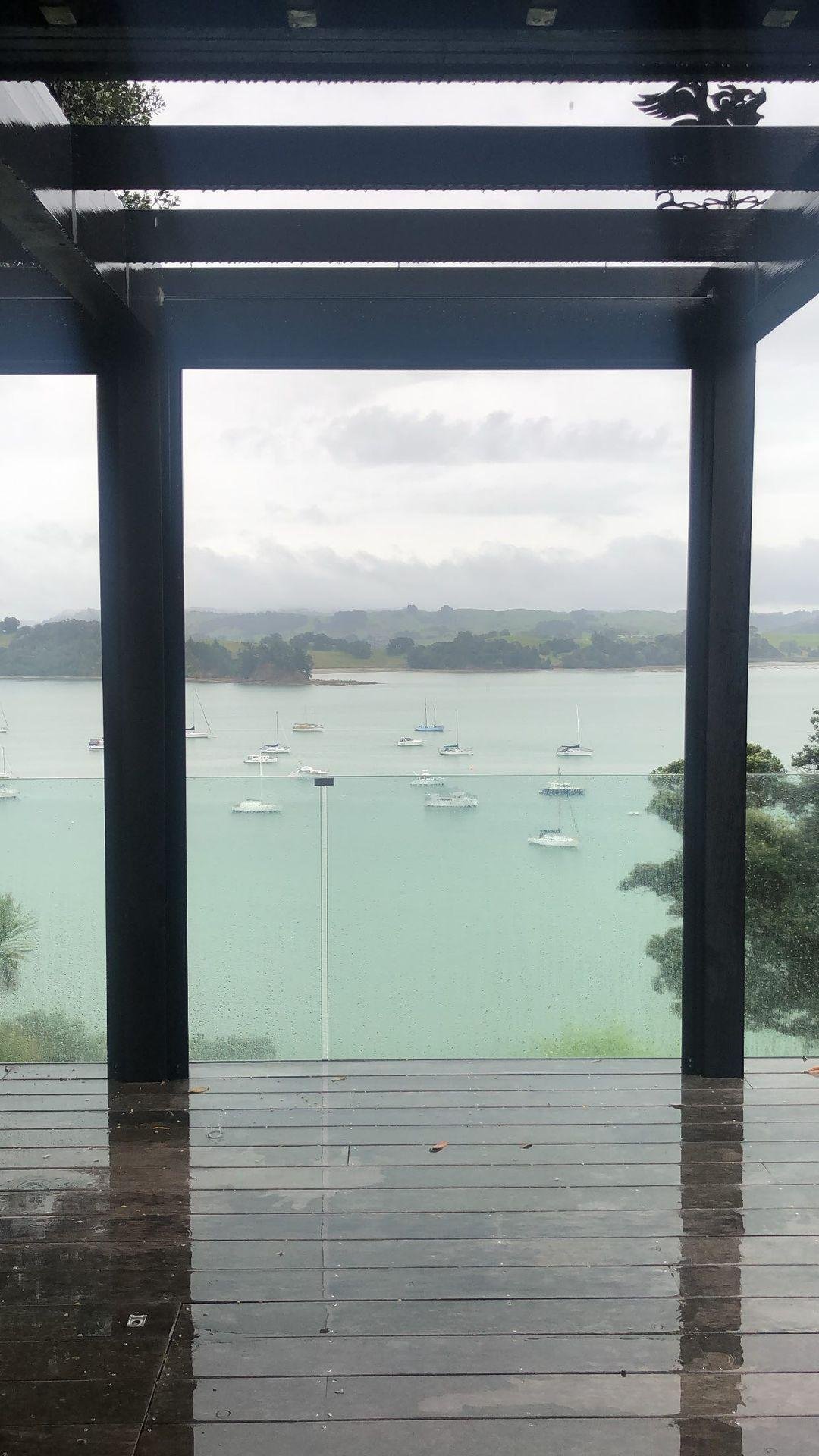 View of a lake with sailboats, seen through a glass railing and framed by a wooden deck and black metal structure on a cloudy day.
