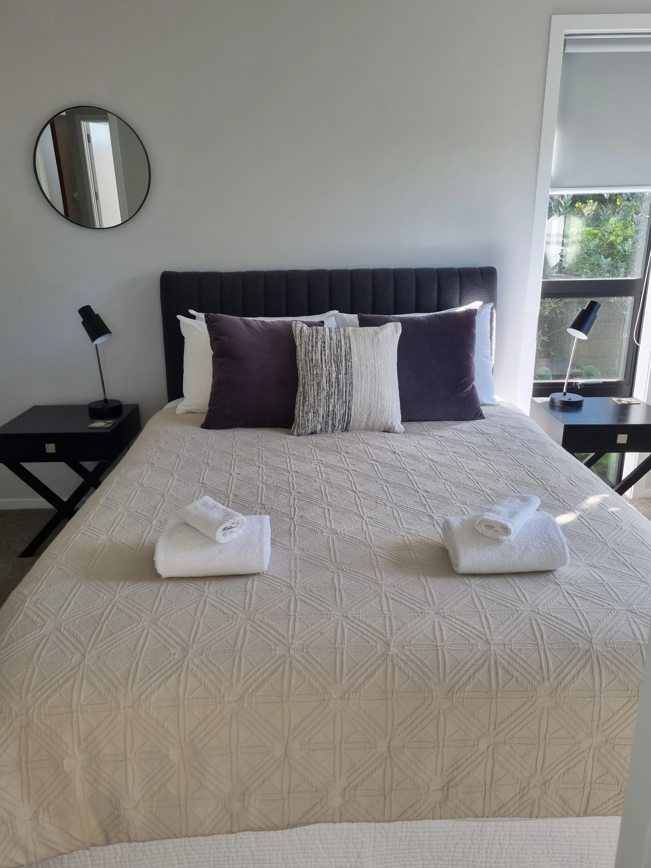 A neatly made bed with cream-colored quilt, with three folded white towels on the foot of the bed. The headboard is black and upholstered. There are decorative pillows, two dark purple and one beige with a textured pattern. Beside the bed, on each side, are black nightstands with black modern desk lamps. A round mirror is on the beige wall, and a window with a partially rolled-up blind and greenery outside is visible on the right.