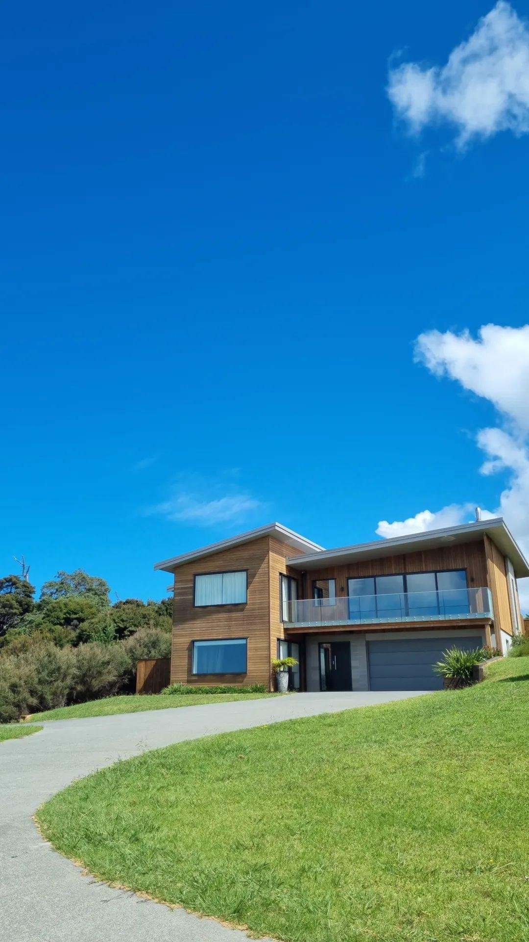 A modern, two-story house with large windows, a wooden exterior, and a spacious balcony. The house is surrounded by green grass and trees, with a bright blue sky and a few white clouds overhead.