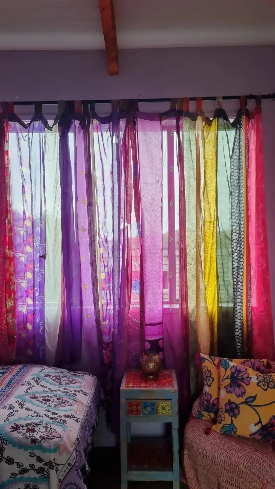 Colorful sheer curtains in purple, pink, green, yellow, and black hanging in front of a window, with patterned bedsheets and cushions visible in the room.