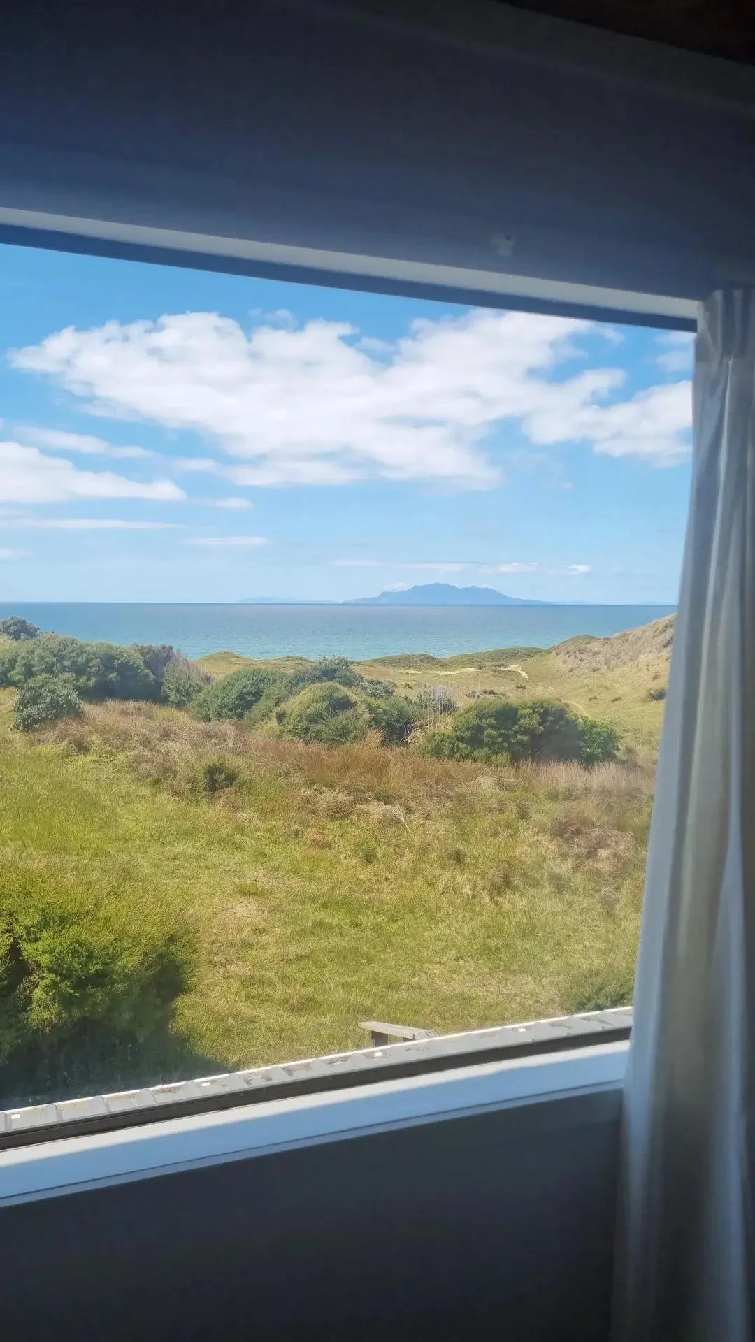 View through a window showing green grassy hills leading to a body of water, with a distant landmass or island on the horizon, and a partly cloudy blue sky.