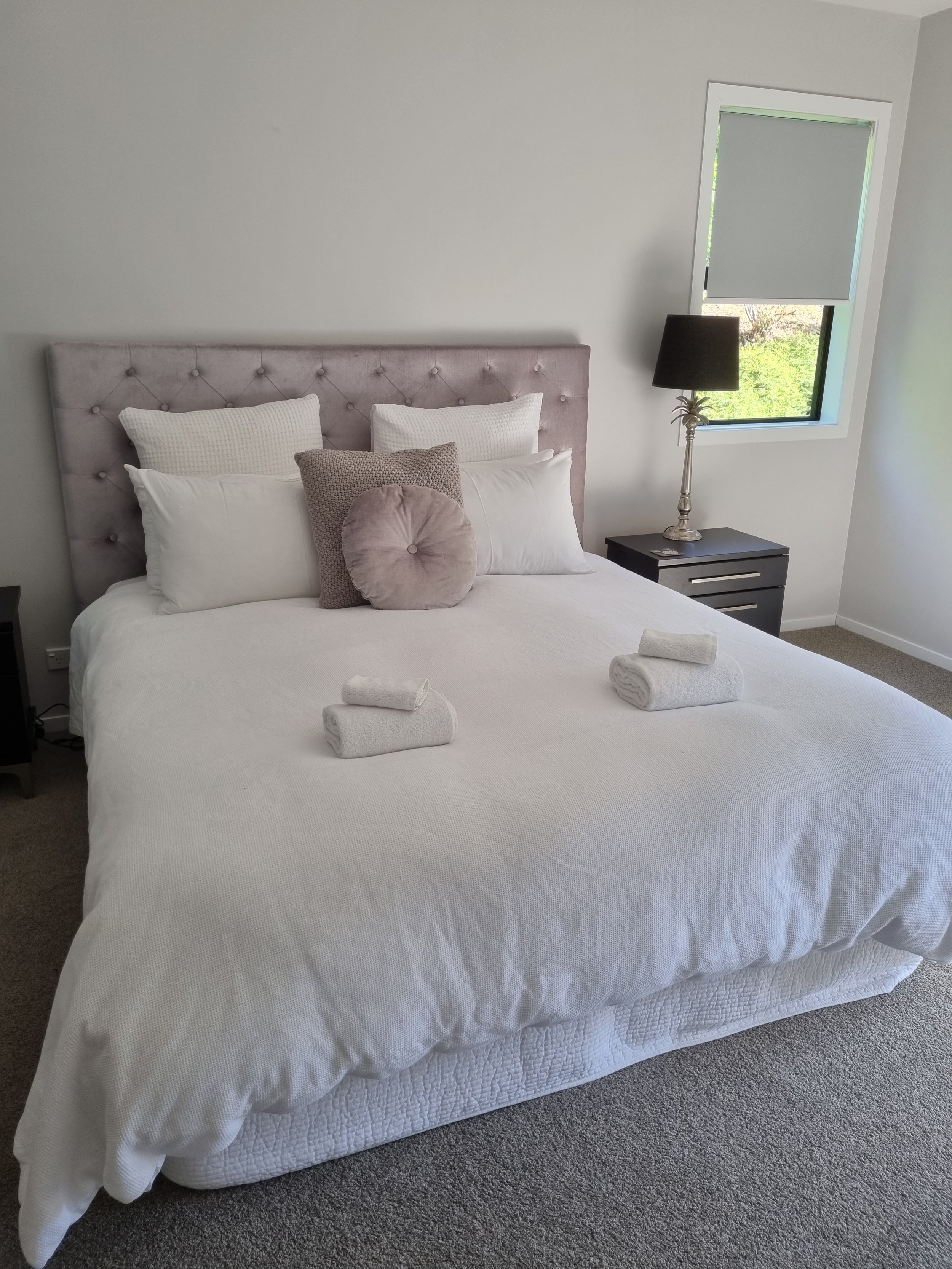 A neatly made bed with white bedding, two large white pillows, a small beige pillow, and a round beige cushion. Two white towels are rolled on the bed. There's a black nightstand with a silver lamp and a remote control. A window with a partially closed blind shows some greenery outside.