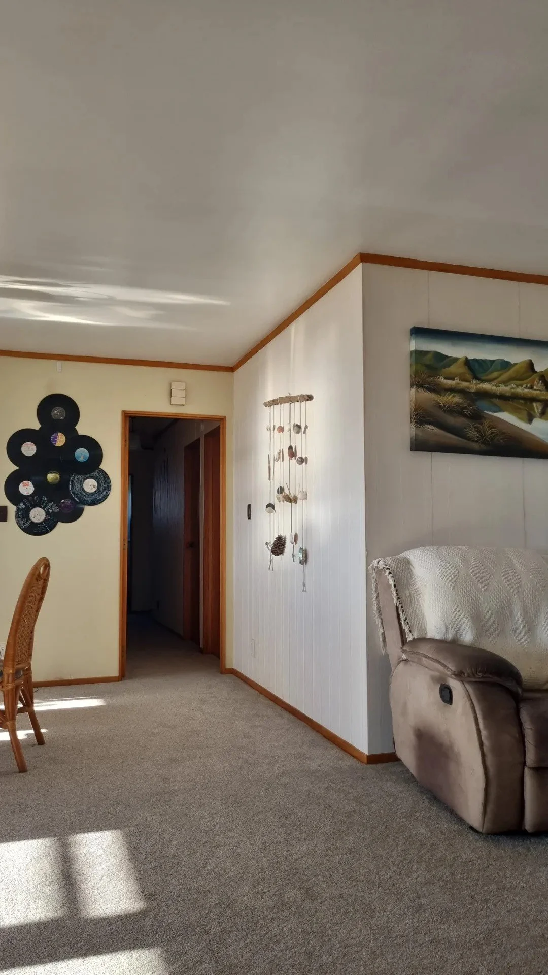 Interior view of a living room with a beige carpet, a corner of a wicker chair, decorative wall hangings including a collection of vinyl records arranged in a circular pattern, a hanging mobile made of seashells and other natural objects, a painting 