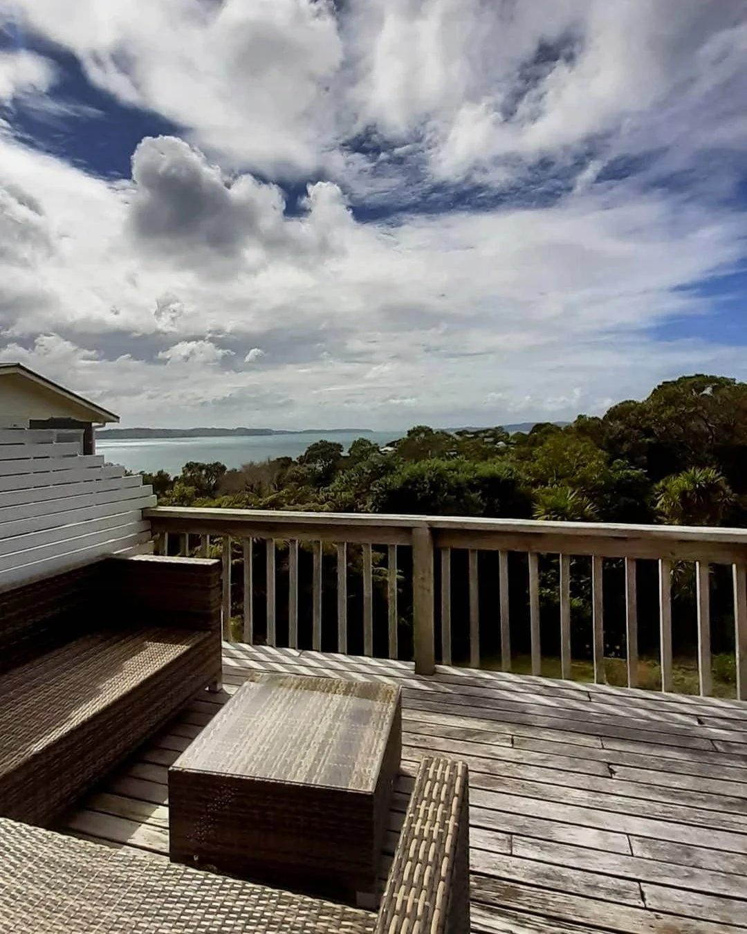 A wooden deck with outdoor furniture overlooking a lush landscape with trees and a body of water in the distance under a partly cloudy sky.