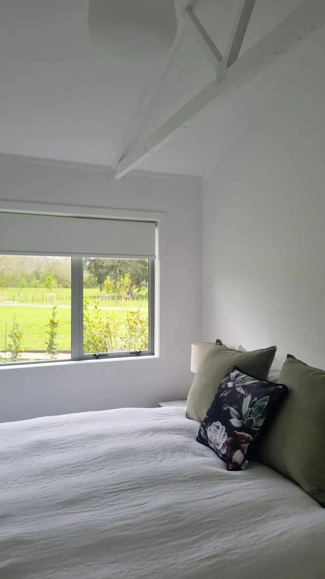 A bedroom with a bed, pillows, a window with a view of greenery outside, and a ceiling vent or beam.
