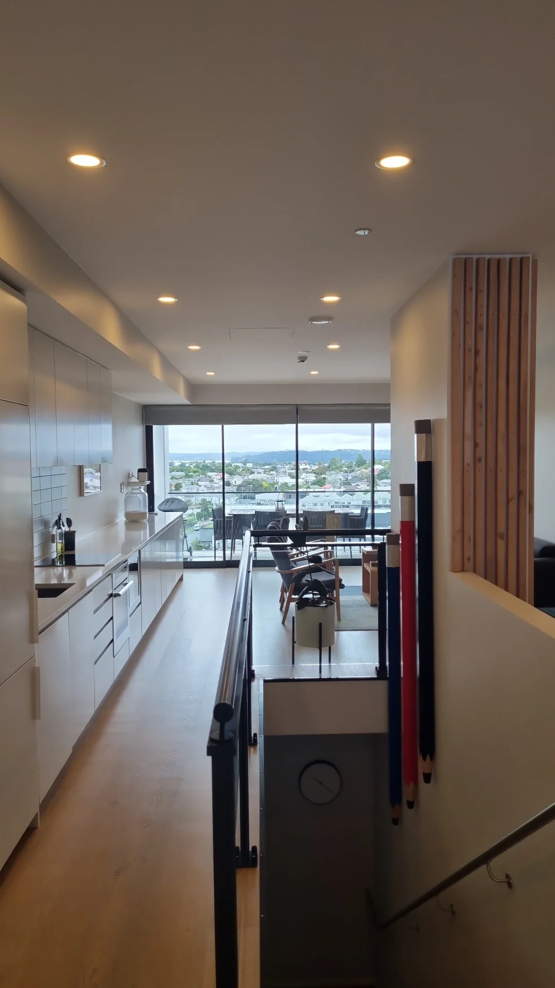 Interior view of a modern apartment with a kitchen on the left, a dining area with chairs, and a large window with a balcony overlooking a cityscape and mountains in the distance.