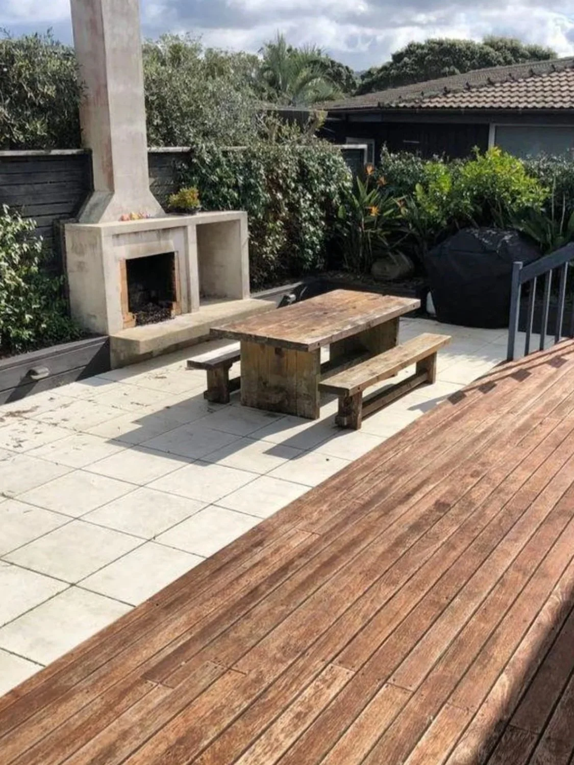 Outdoor patio with a rustic wooden picnic table and benches, a white brick fireplace, surrounded by greenery and a wooden fence, with a house roof in the background.