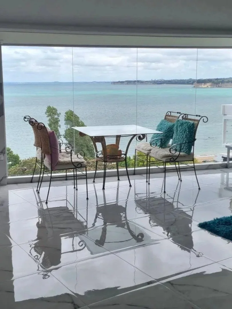 A glass table with four wrought iron chairs with cushions, overlooking a large body of water and distant coastline.
