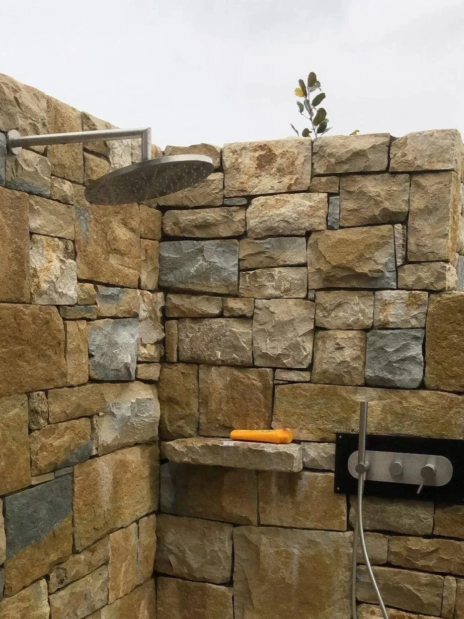 Outdoor stone shower with a round showerhead, a small shelf with a yellow handle tool, and a black panel with controls, built with beige and gray stones.