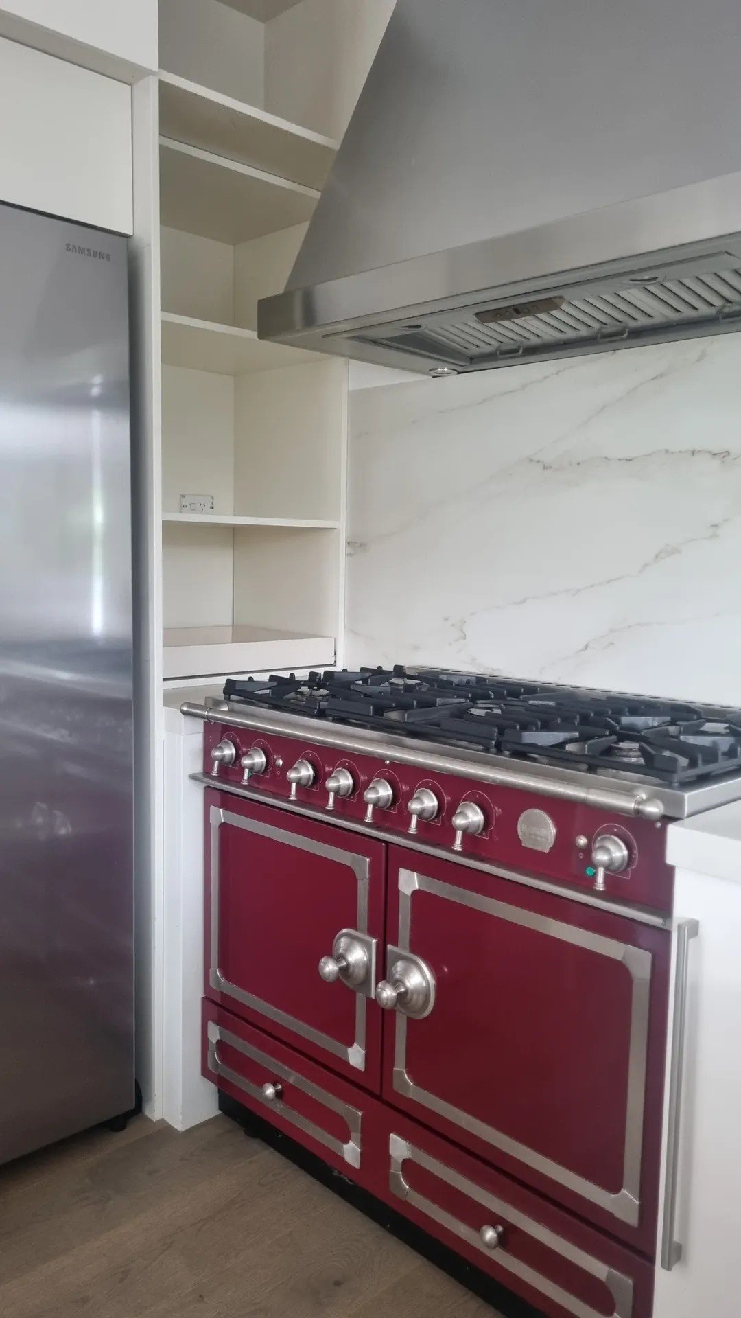 Red vintage-style gas stove with silver knobs and two ovens, next to a silver refrigerator in a modern kitchen with white shelves and marble backsplash.