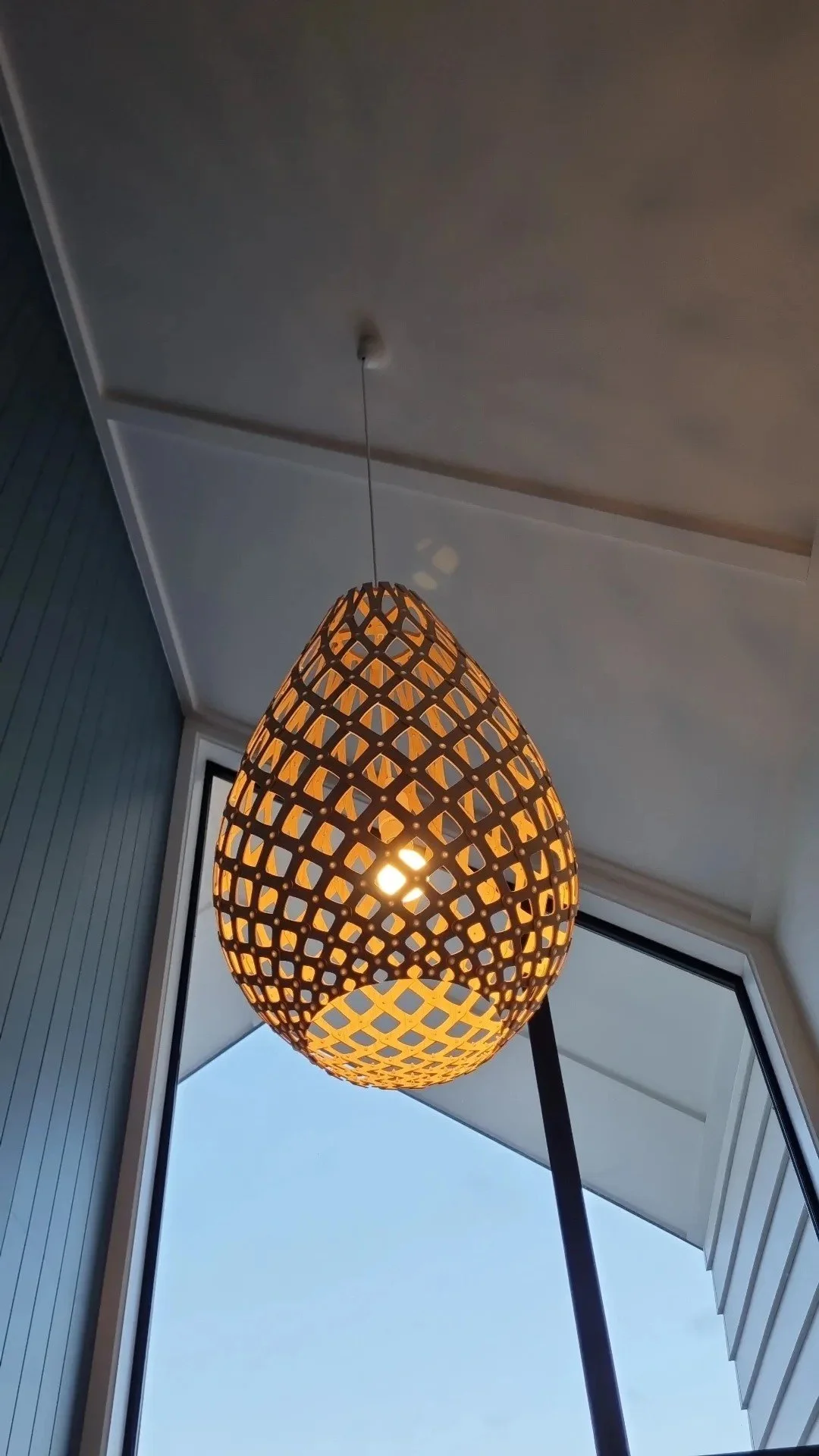 A decorative hanging pendant light with a woven lattice design, emits a warm glow near a large window with a view of blue sky.