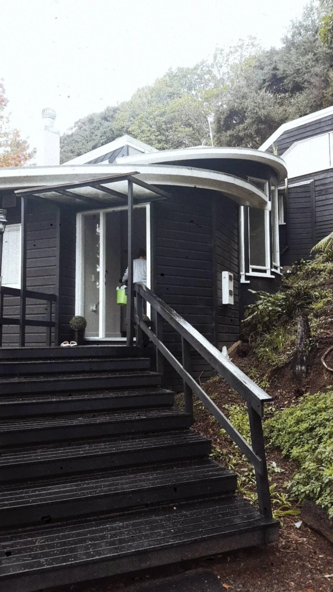 A black house with a black wooden staircase leading to the front door, which is open. There is a person standing at the door. The house has a small porch area with a green mailbox attached to the wall. Surrounding the house are trees and plants.