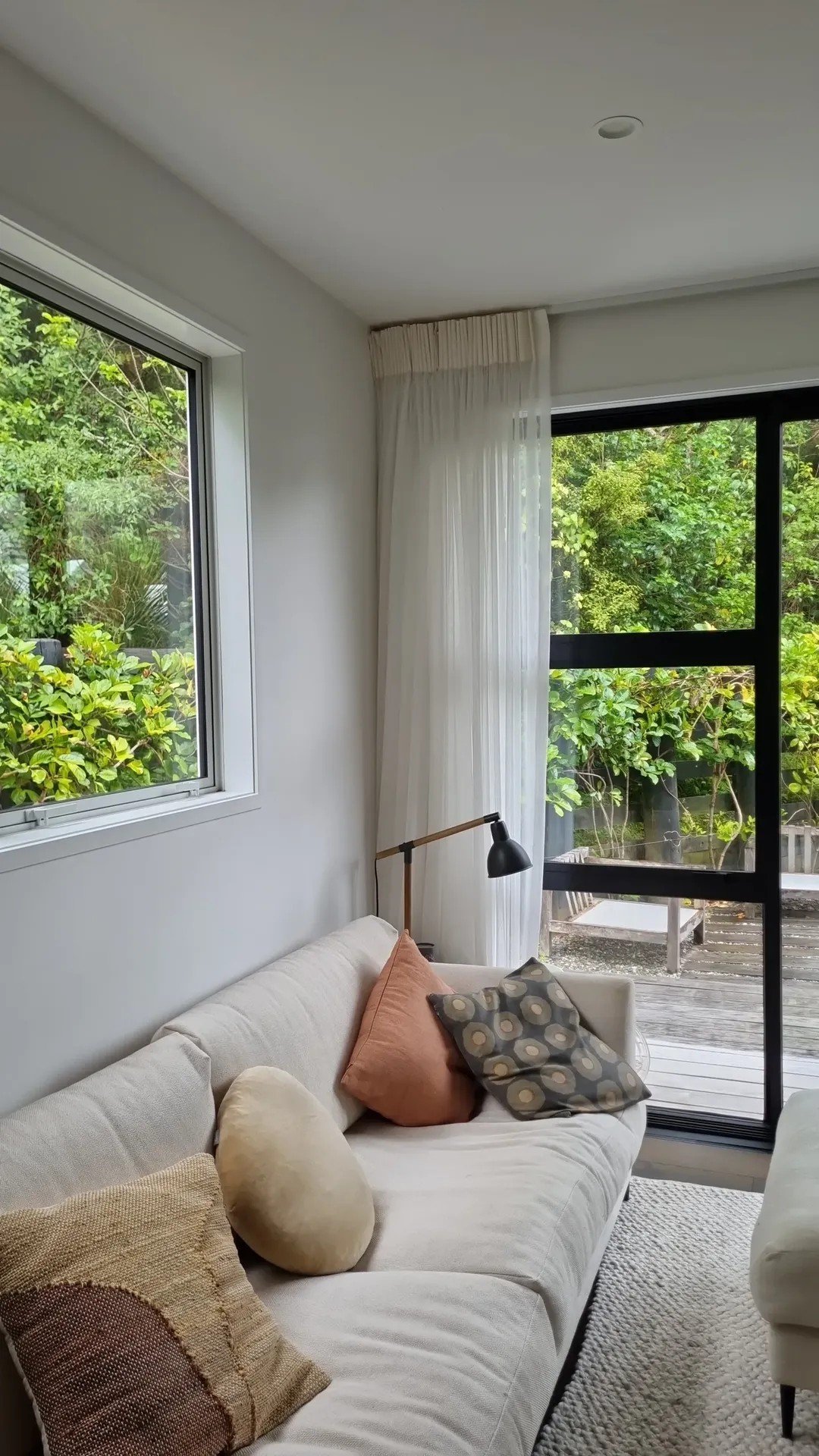 Living room with white sofa, assorted pillows, black side lamp, large window showing greenery outside, white curtains, and sliding glass door leading to an outdoor deck.