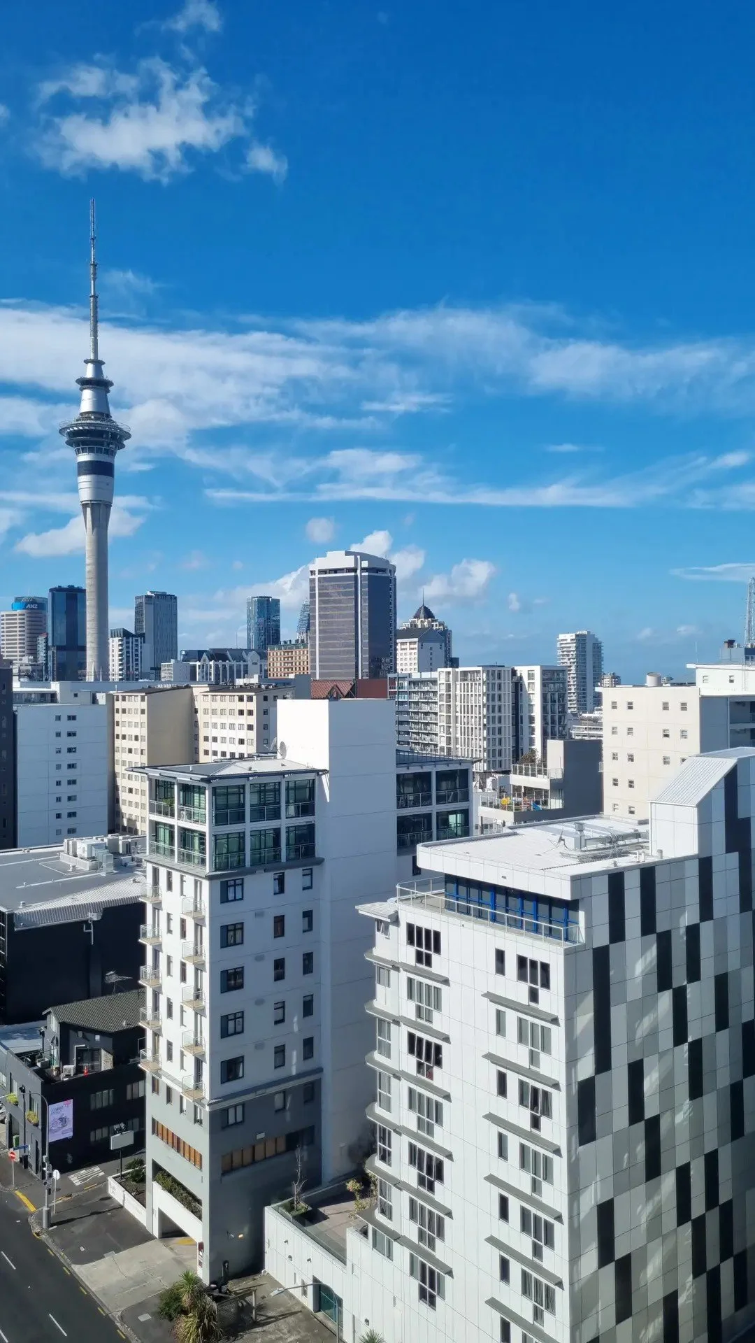 City skyline with modern white apartment buildings in foreground and tall skyscrapers, including the Sky Tower, against a partly cloudy blue sky in the background.