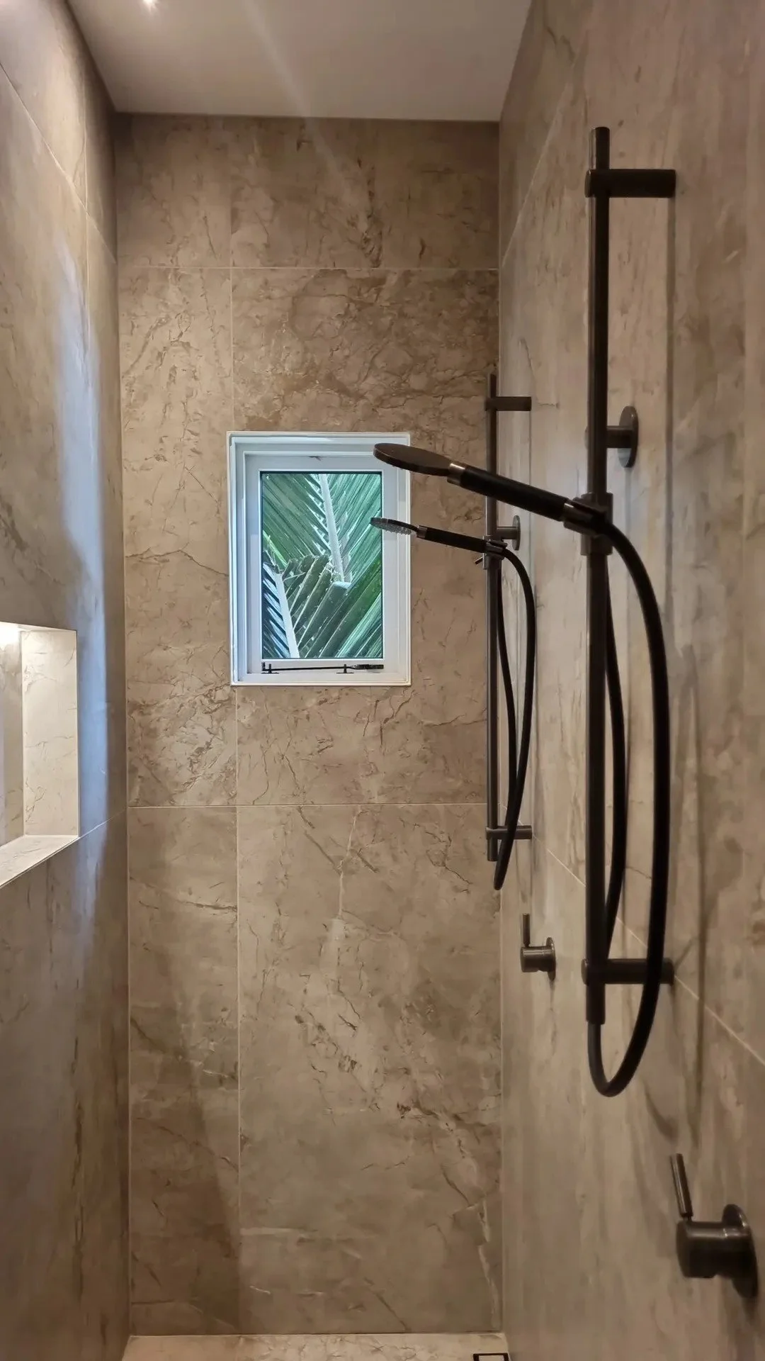 View of a walk-in shower with beige marble tiles, a small window with green plants outside, and black shower fixtures including a showerhead, a handheld sprayer, and a water control knob.